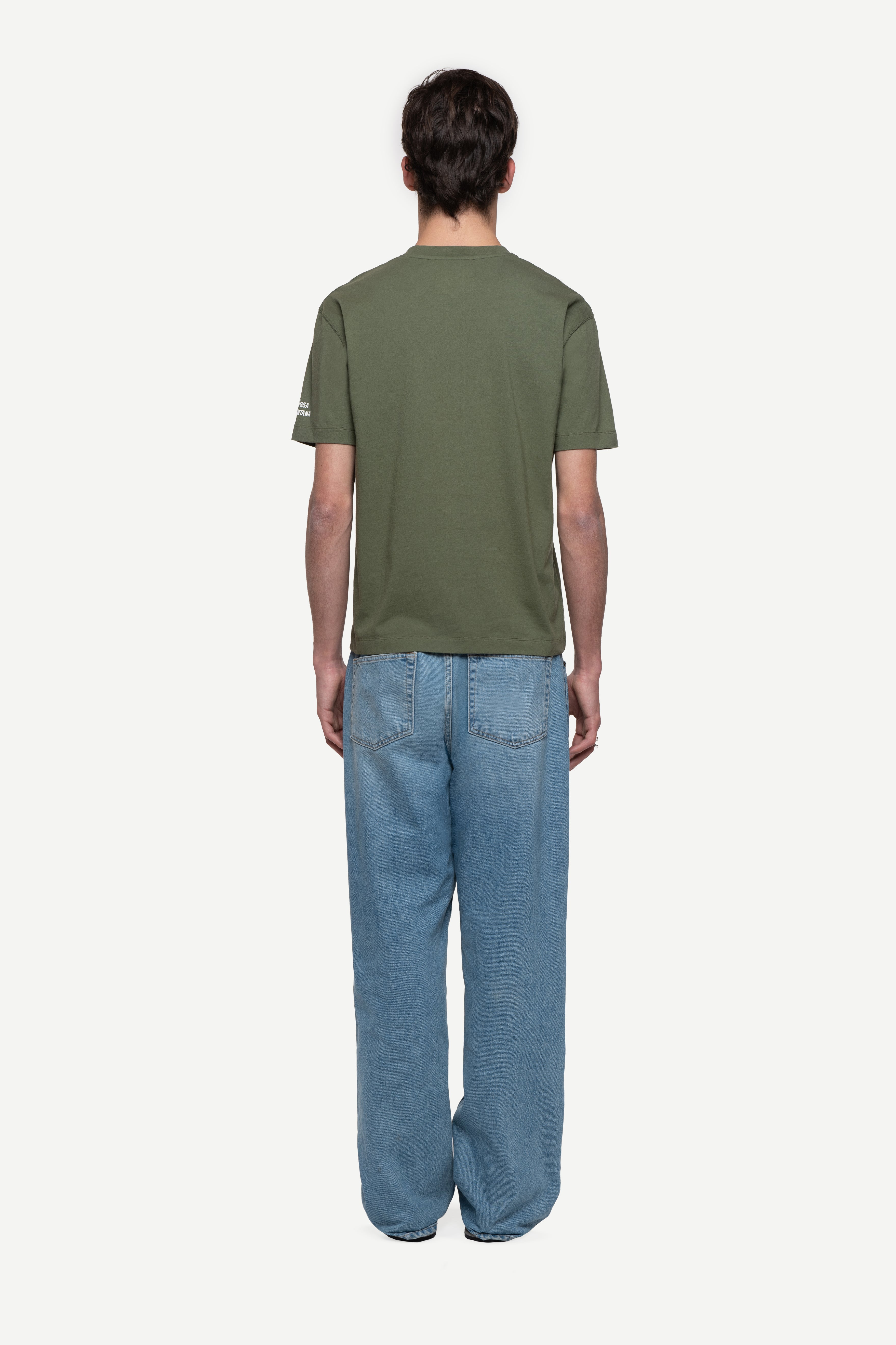 back view of man wearing a regular fit green short-sleeved t-shirt, blue jeans and flip flops on a white background