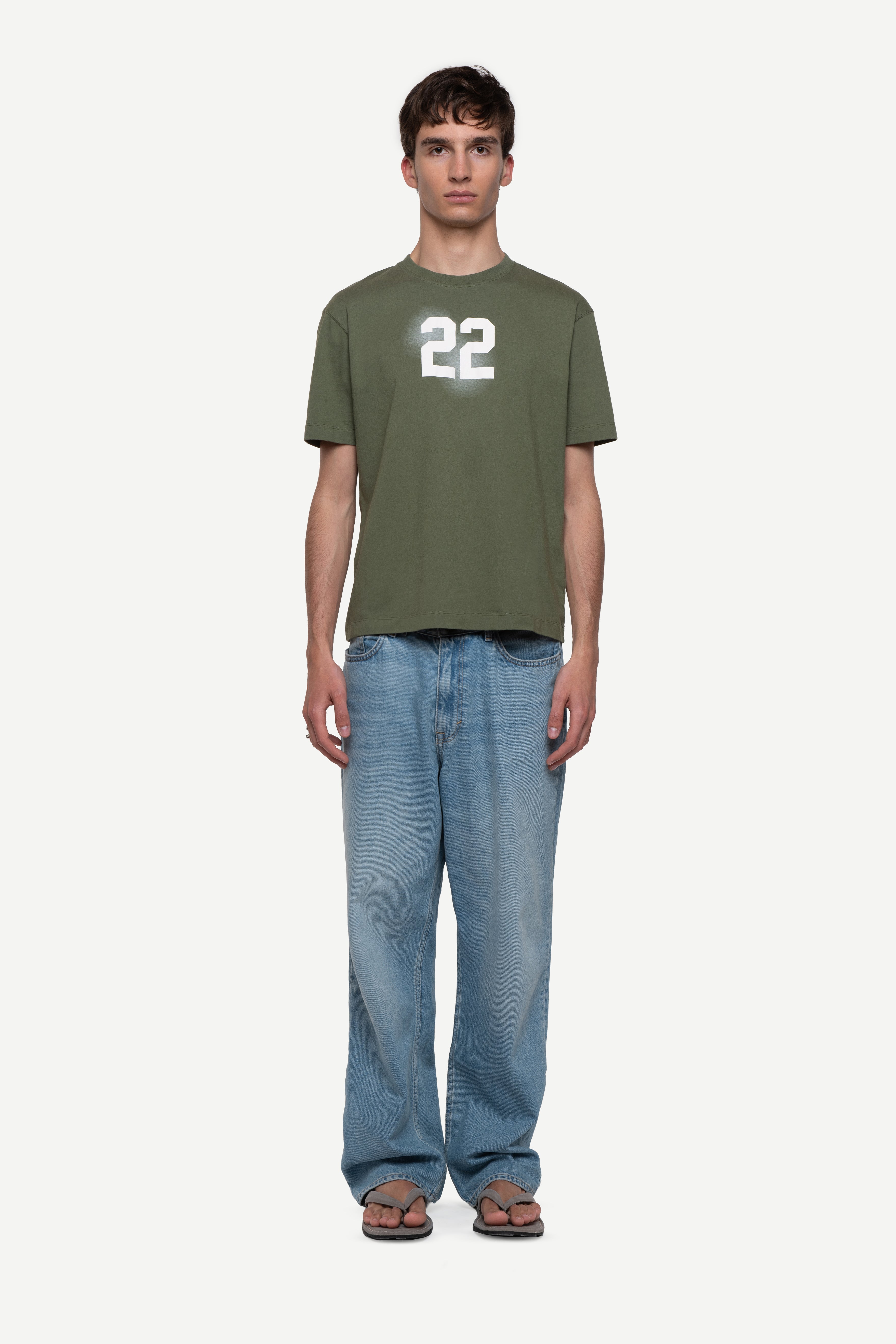 front view of man wearing a regular fit green short-sleeved t-shirt with ivory cloudy "22" print on the chest and "demôssa, tramuntana" sleeve print, blue jeans and flip flops on a white background