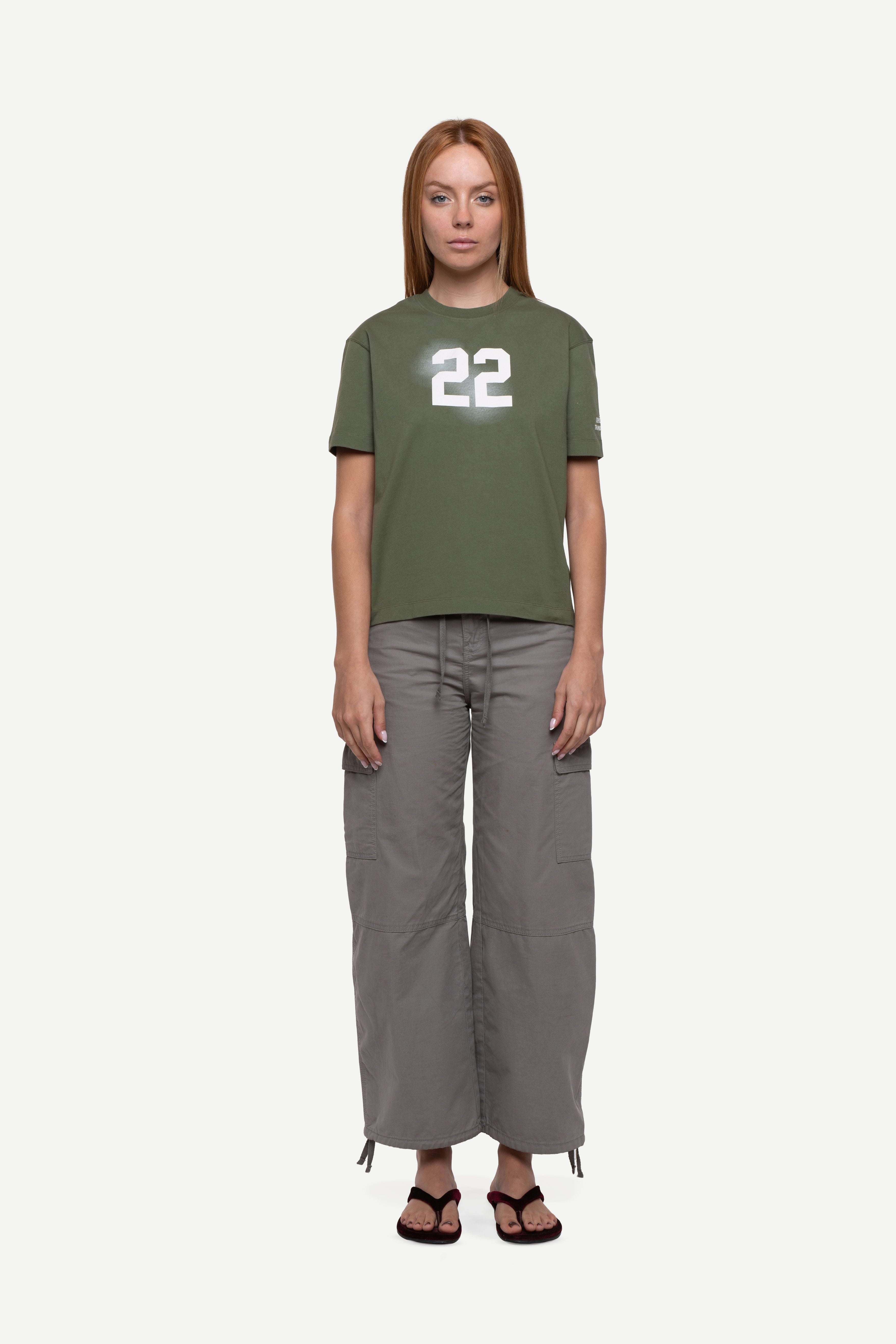 front view of woman wearing a regular fit green short-sleeved t-shirt with ivory cloudy "22" print on the chest and "demôssa, tramuntana" sleeve print, grey cargo pants and flip flops on a white background