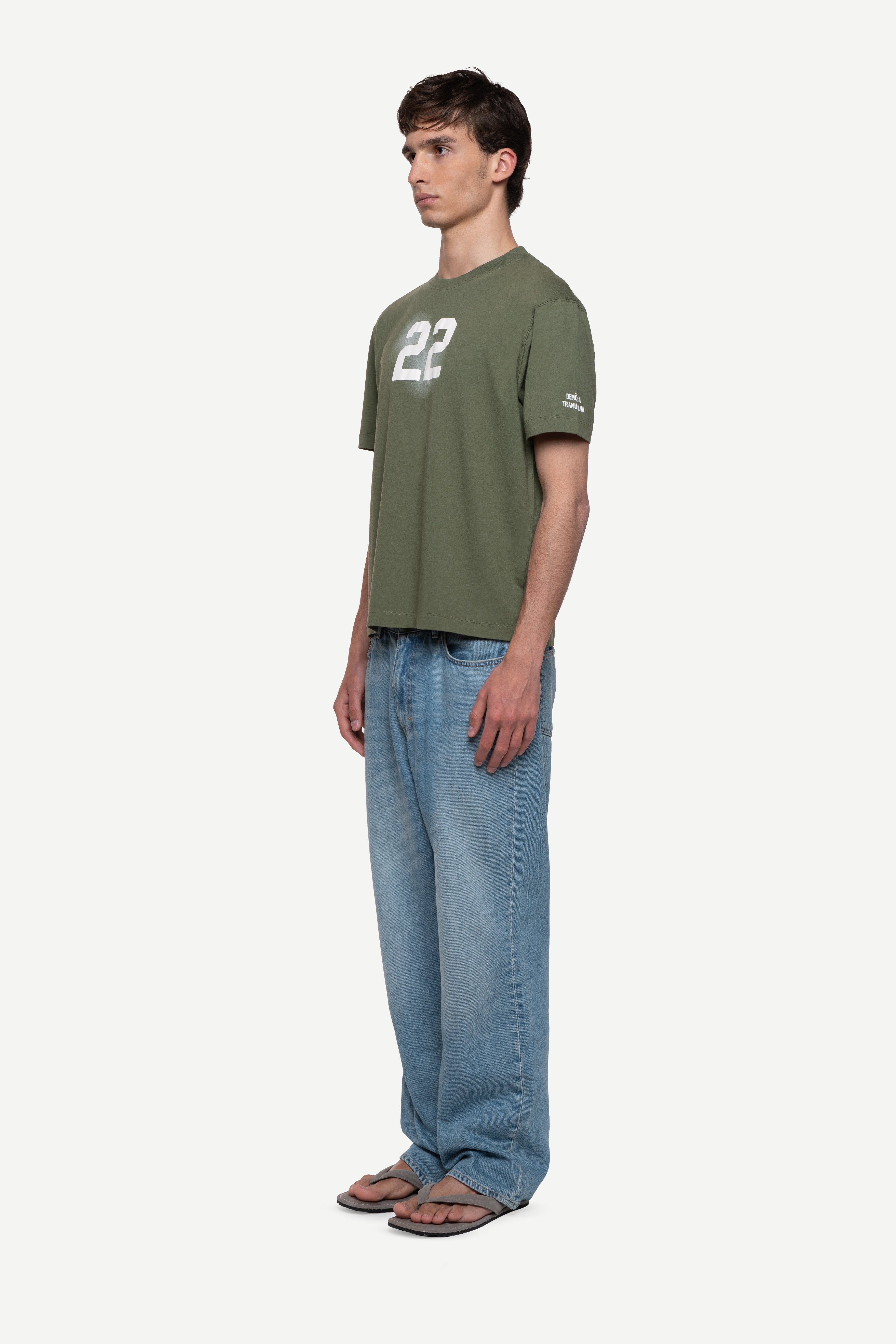 side view of man wearing a regular fit green short-sleeved t-shirt with ivory cloudy "22" print on the chest and "demôssa, tramuntana" sleeve print, blue jeans and flip flops on a white background