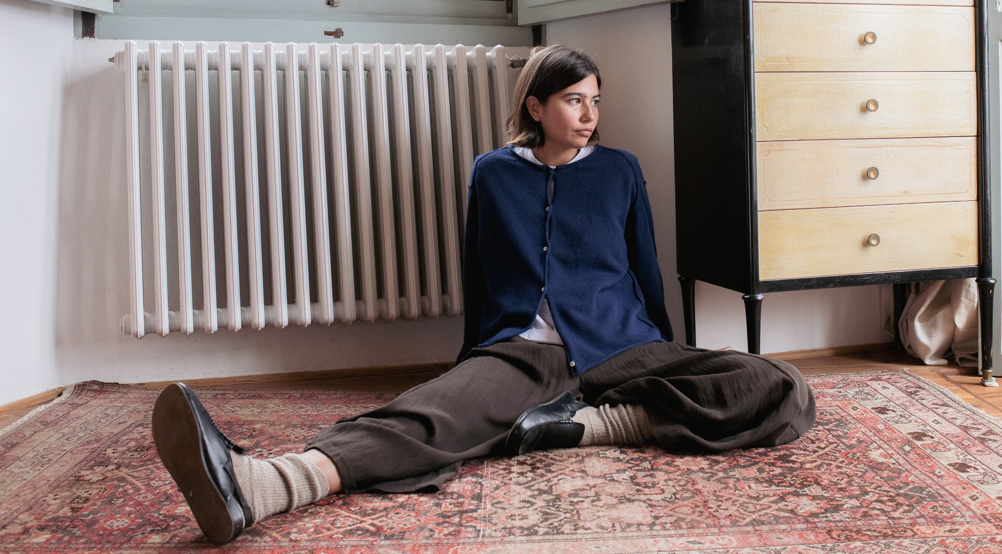 Person sitting on a patterned rug in a room with a radiator and wooden furniture.
