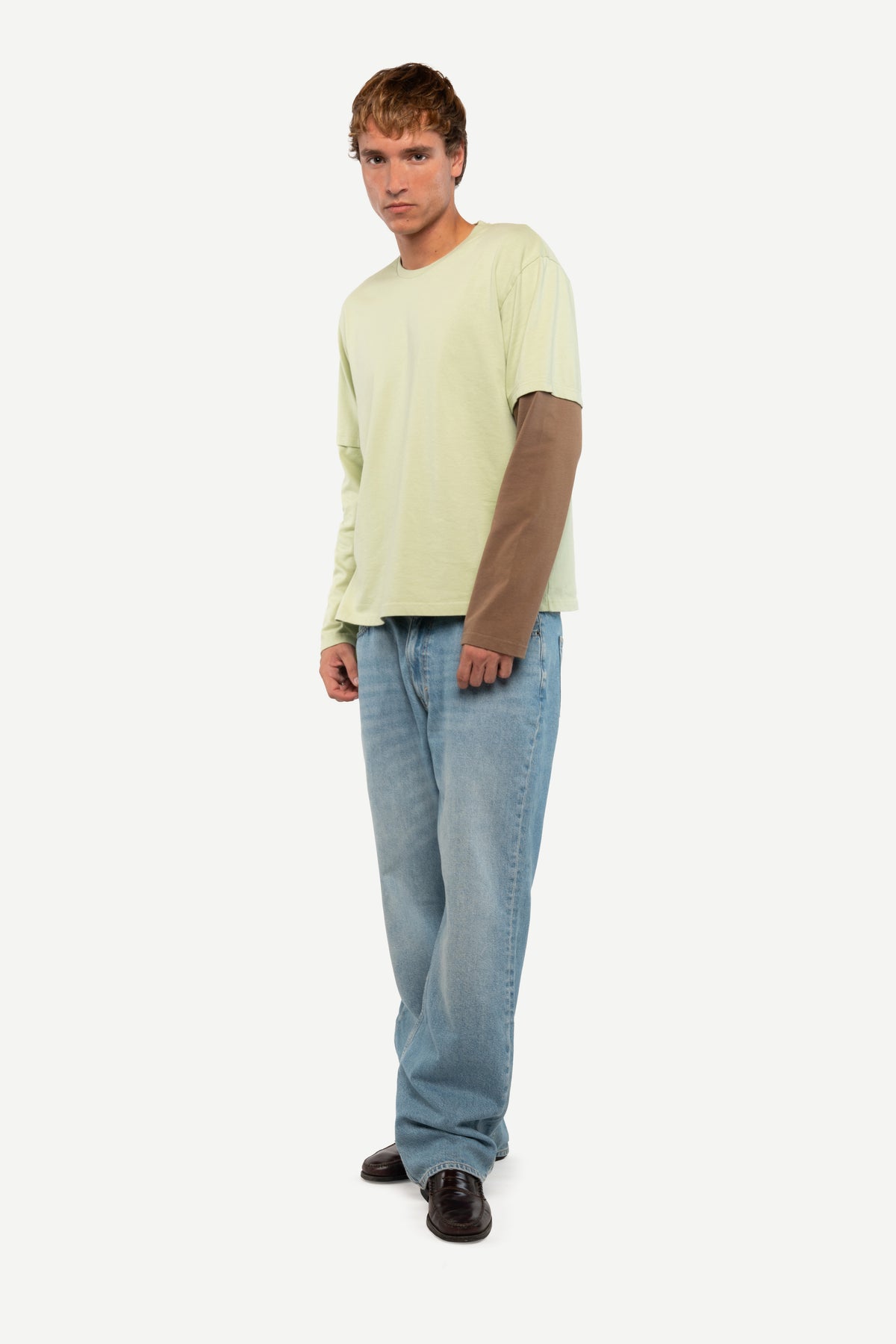 A man modeling a light yellow-green long-sleeve t-shirt layered over a brown long-sleeve shirt, paired with light blue jeans and dark brown shoes, against a plain light gray background.
