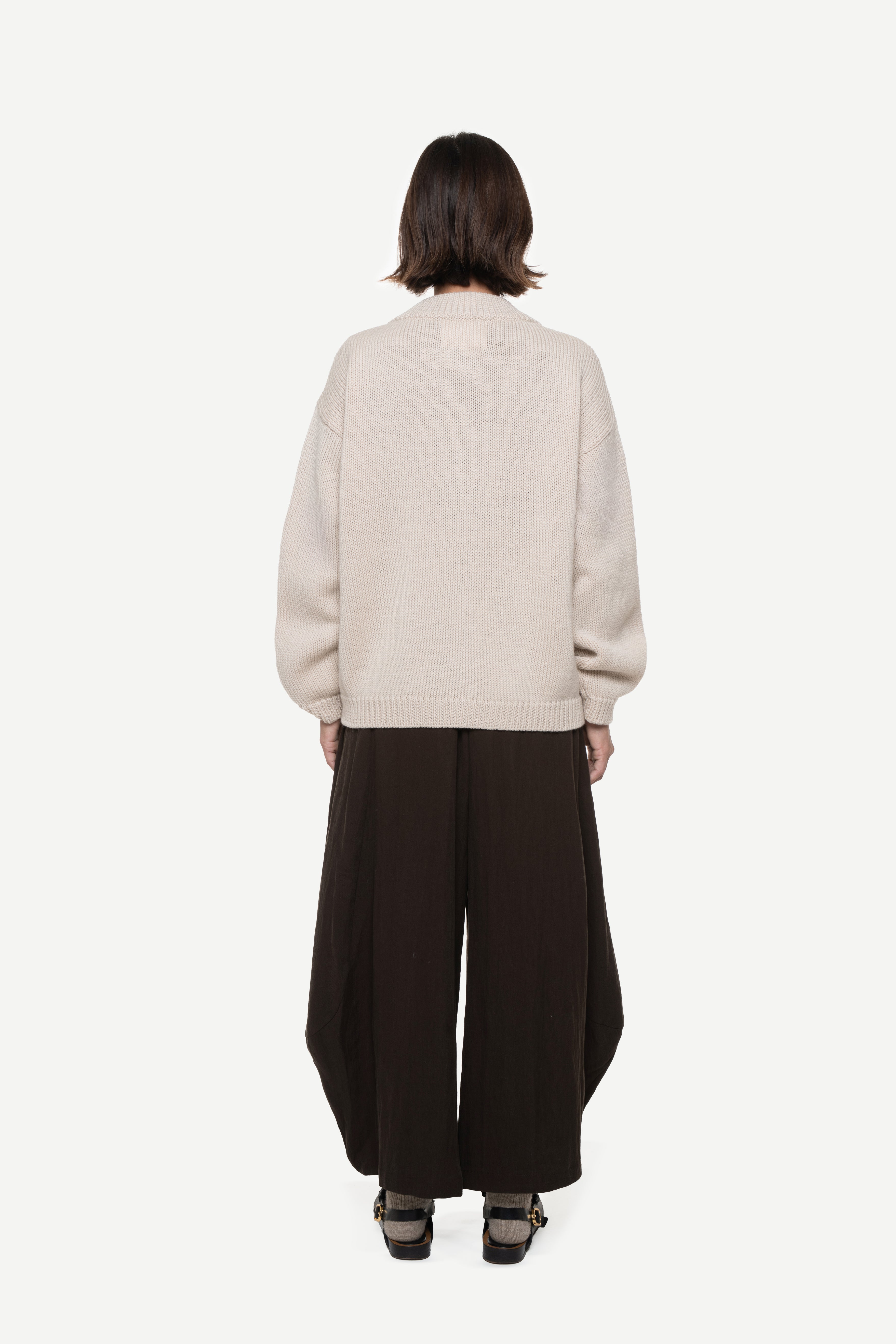 back view of a woman wearing a beige cardigan and brown pants on a white background