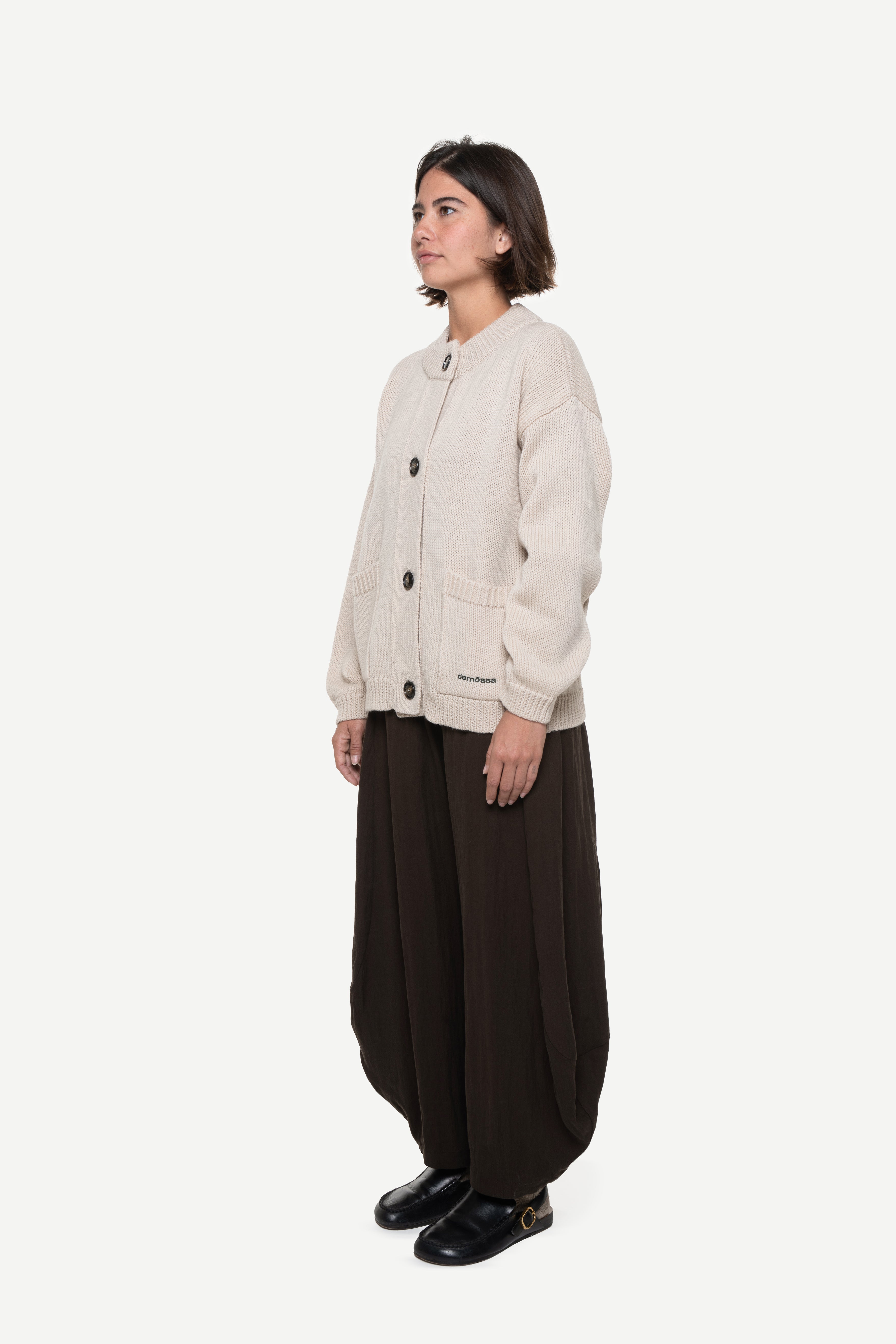 side view of a woman wearing a beige buttoned cardigan with two front pockets and brown pants on a white background