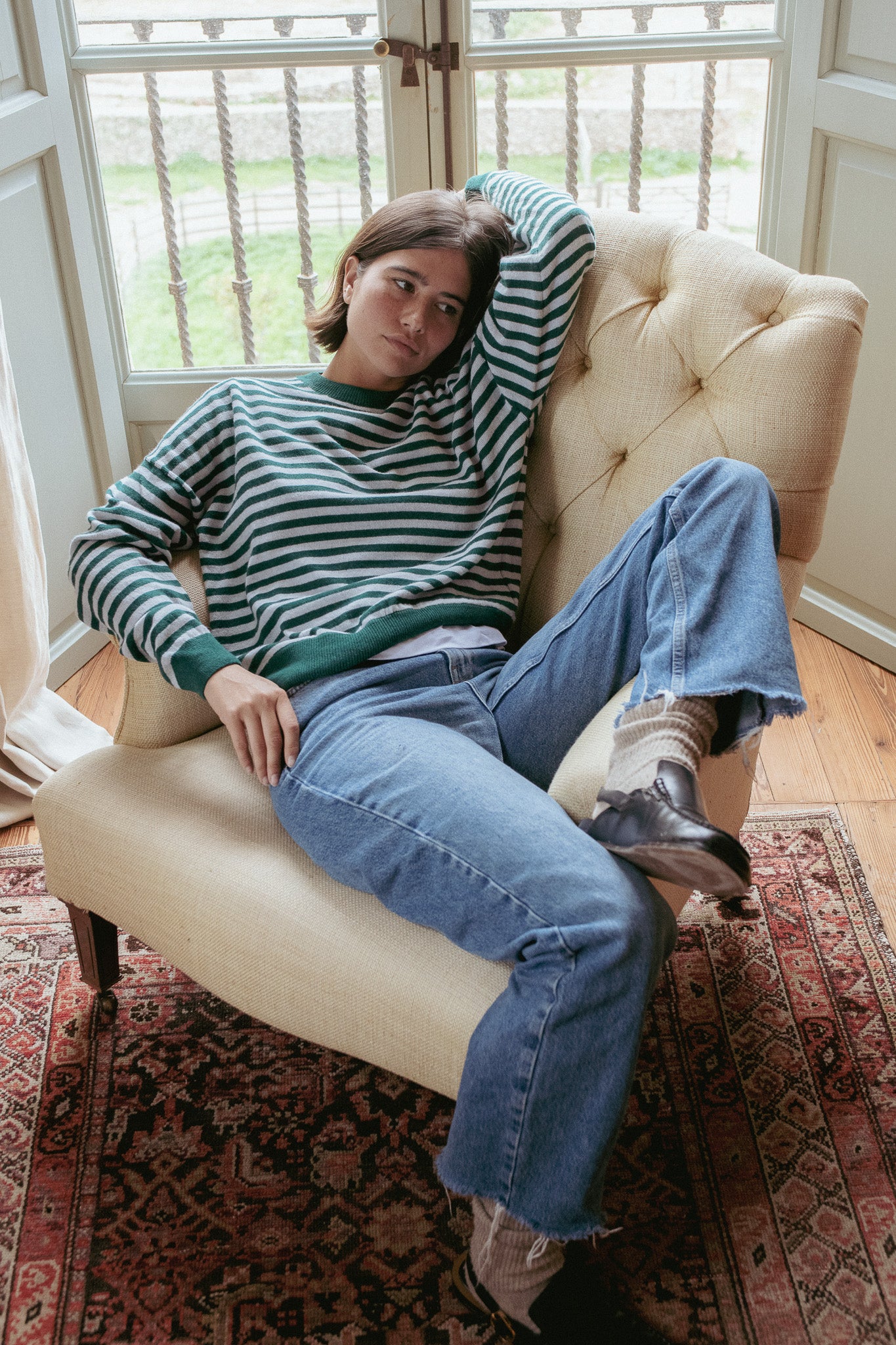 Person sitting on a beige armchair wearing a striped sweater and jeans in a room with large windows.
