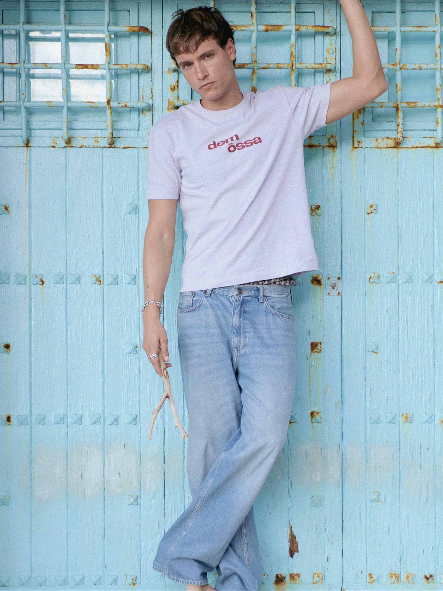 Person wearing a light gray t-shirt with red text and light blue jeans against a blue wooden gate.