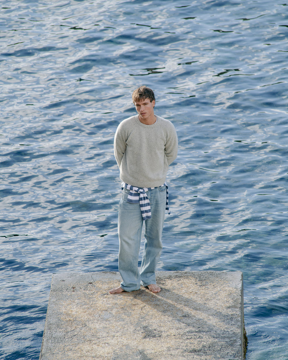 Man standing on a dock by the water wearing a light gray sweater and blue jeans.
