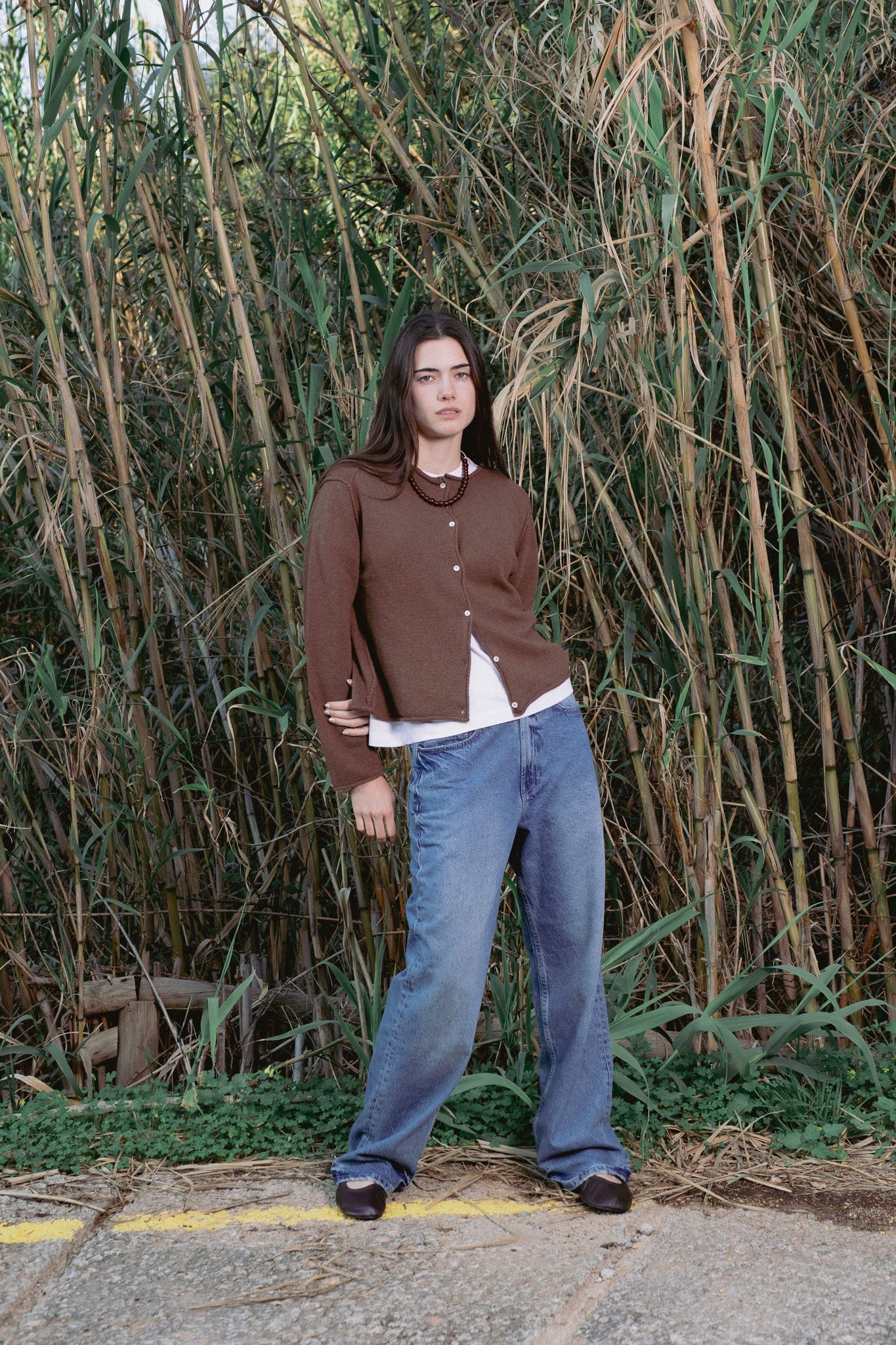Person wearing a brown cardigan and blue jeans standing in front of tall grass.