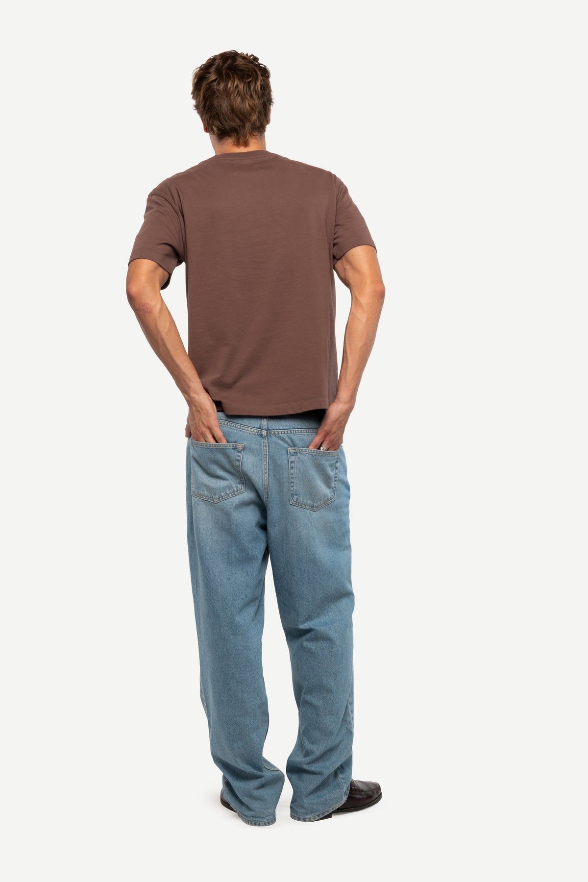 A man viewed from behind wearing a brown short-sleeve t-shirt and light blue jeans, standing against a plain light gray background.