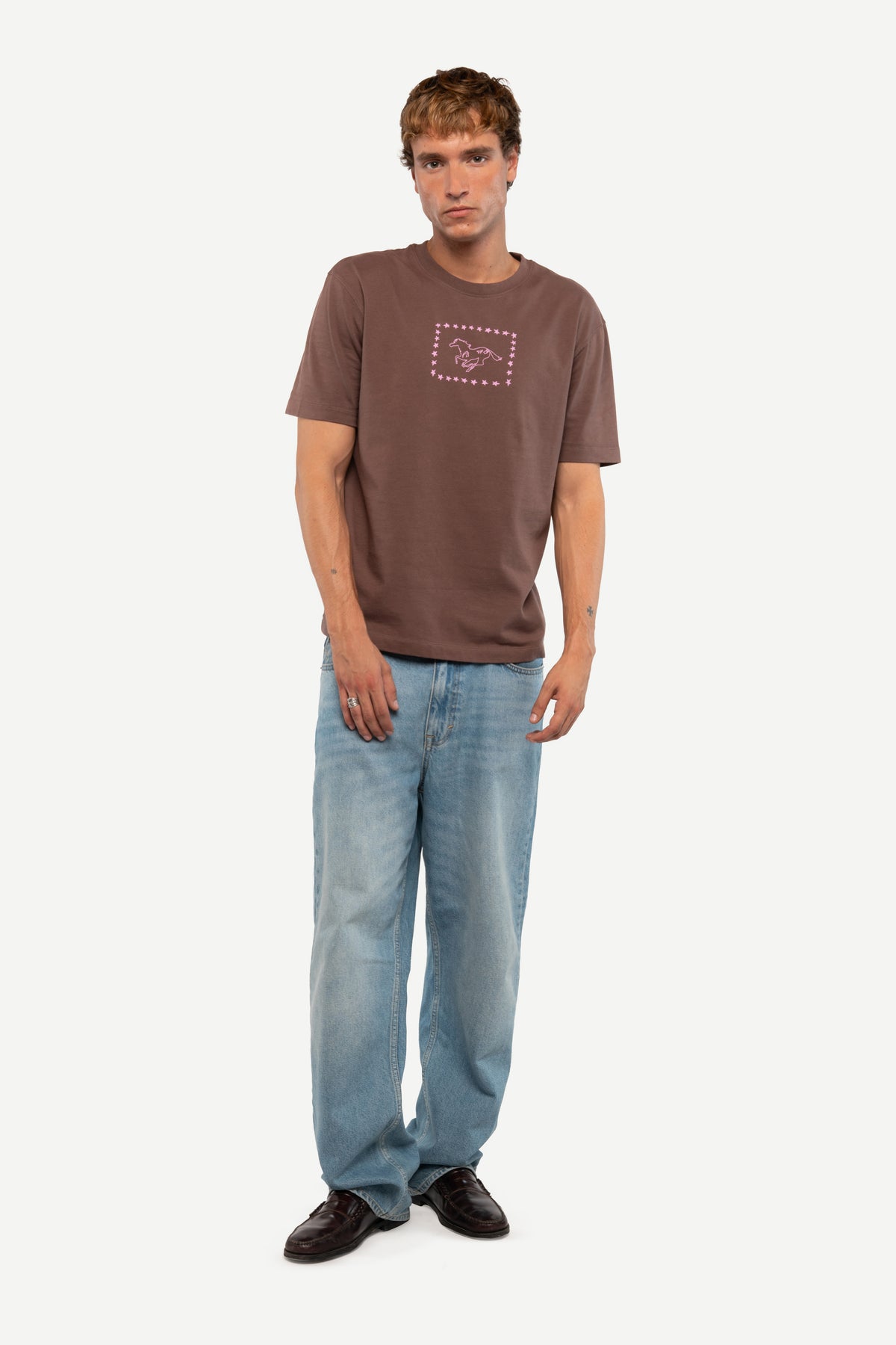 A man modeling a brown short-sleeve t-shirt with a small graphic on the chest, paired with light blue jeans and black shoes, against a plain light gray background.