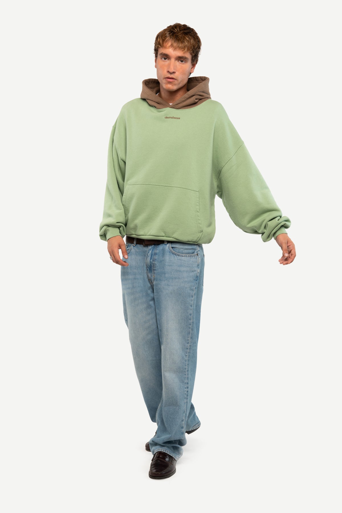 A man modeling a light green oversized hoodie with a contrasting brown hood lining, paired with light blue jeans and dark brown shoes, against a plain light gray background.