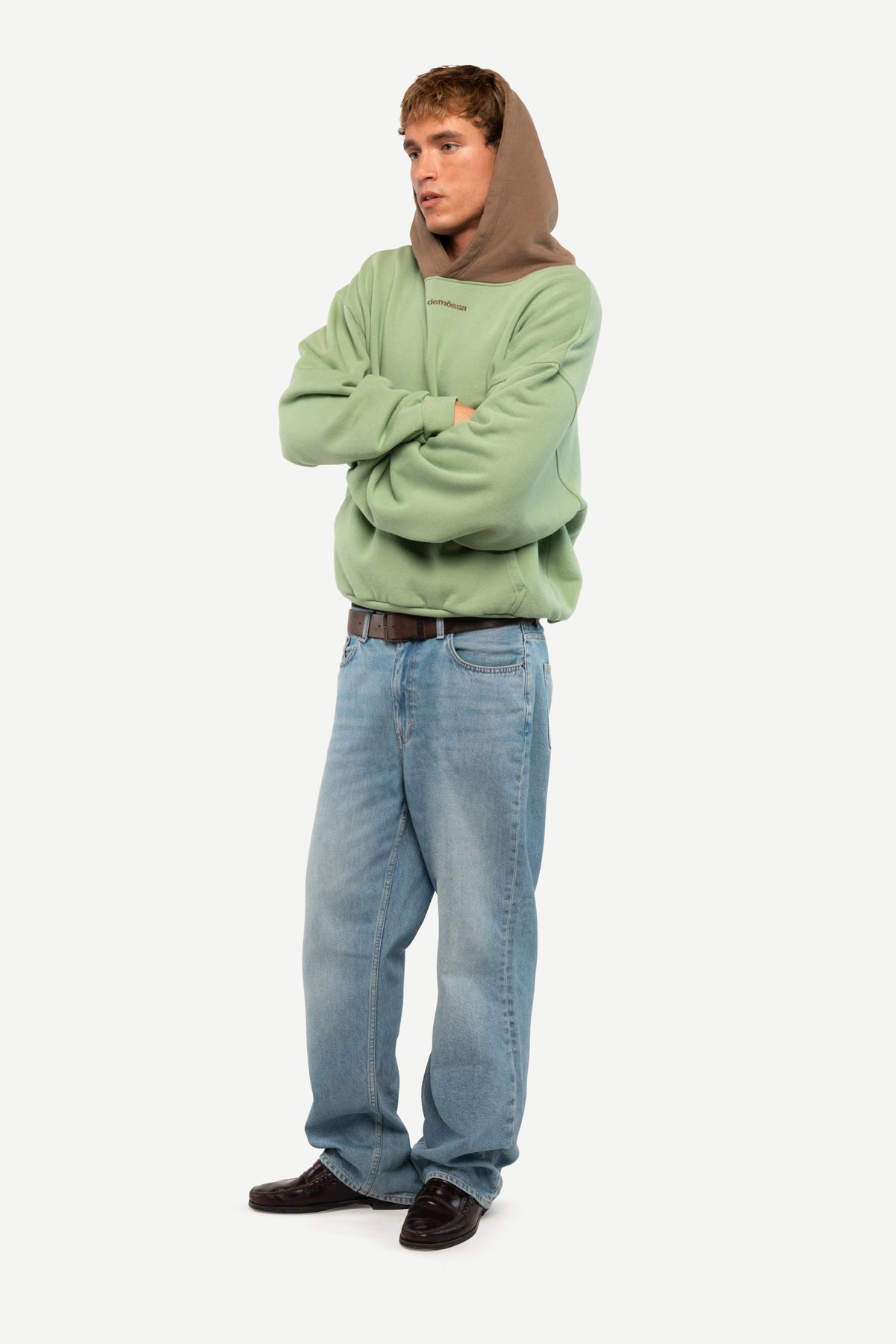 A man modeling a sage green hoodie with a brown hood lining, paired with light blue jeans and dark brown shoes, standing against a plain light gray background.