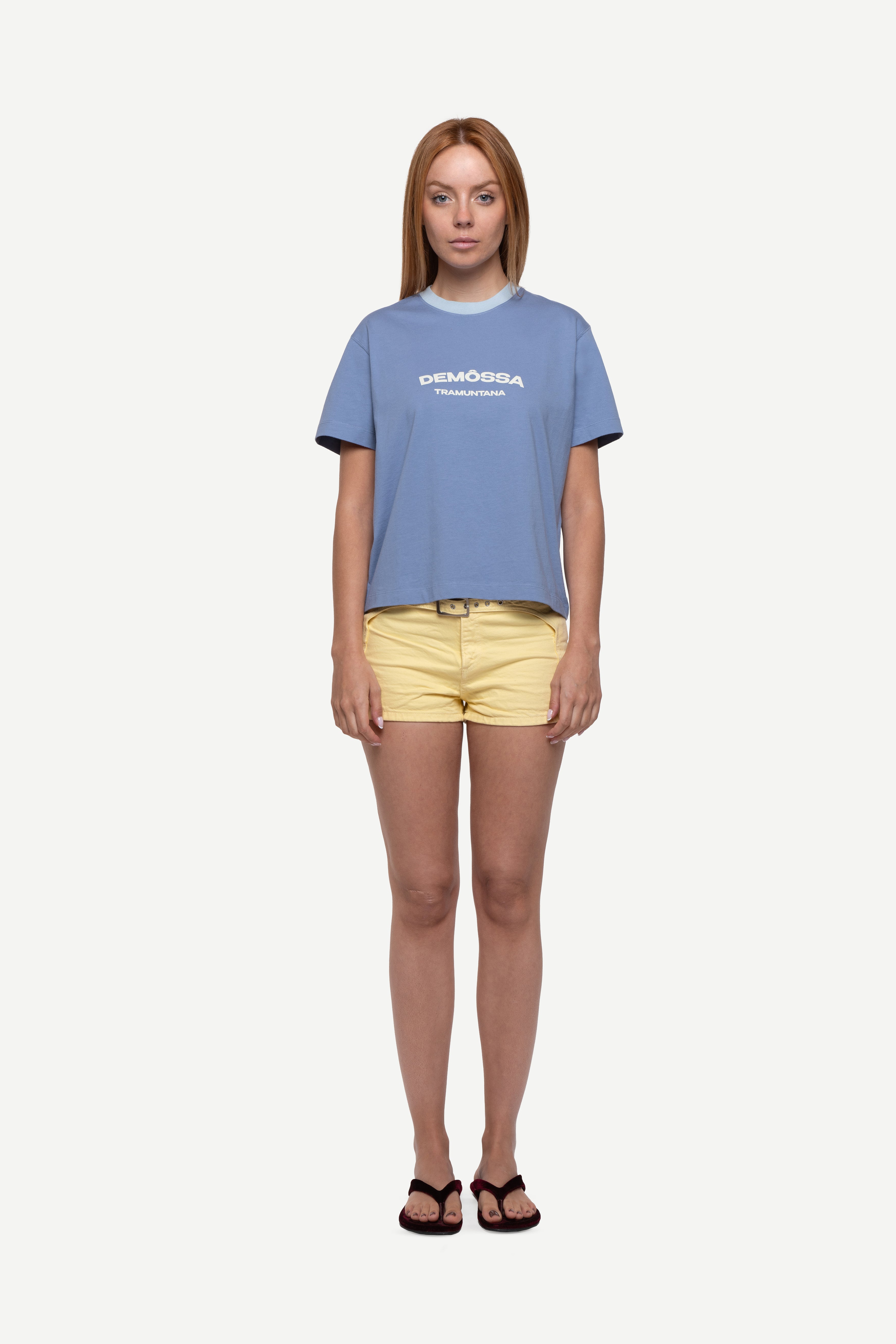 front view of woman wearing a regular fit blue short-sleeved t-shirt with ivory demôssa print on the chest, yellow shorts and flip flops on a white background