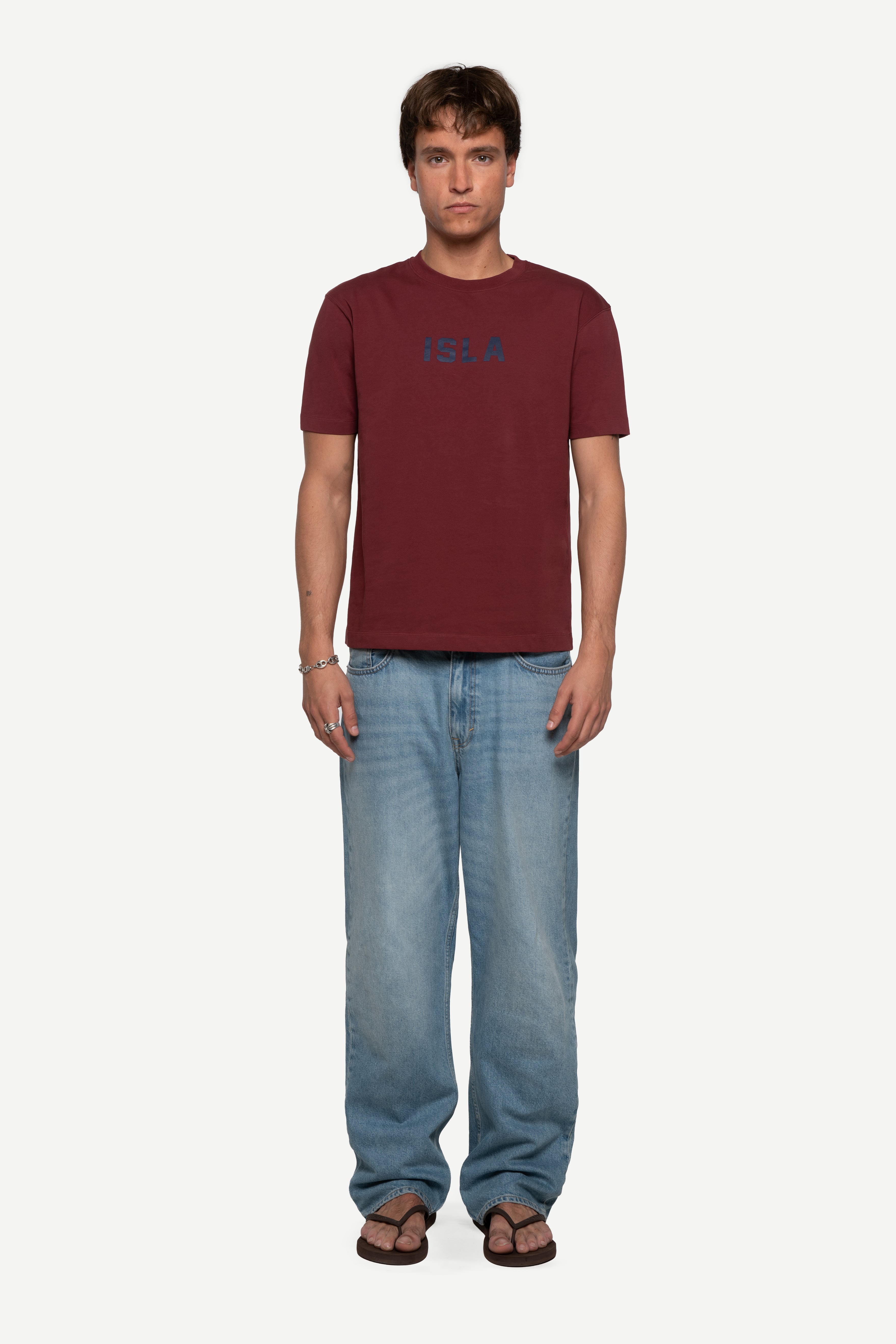 front view of man wearing a regular fit short-sleeved burgundy t-shirt featuring a tonal 'demôssa' logo print on the chest with blue jeans on a white background