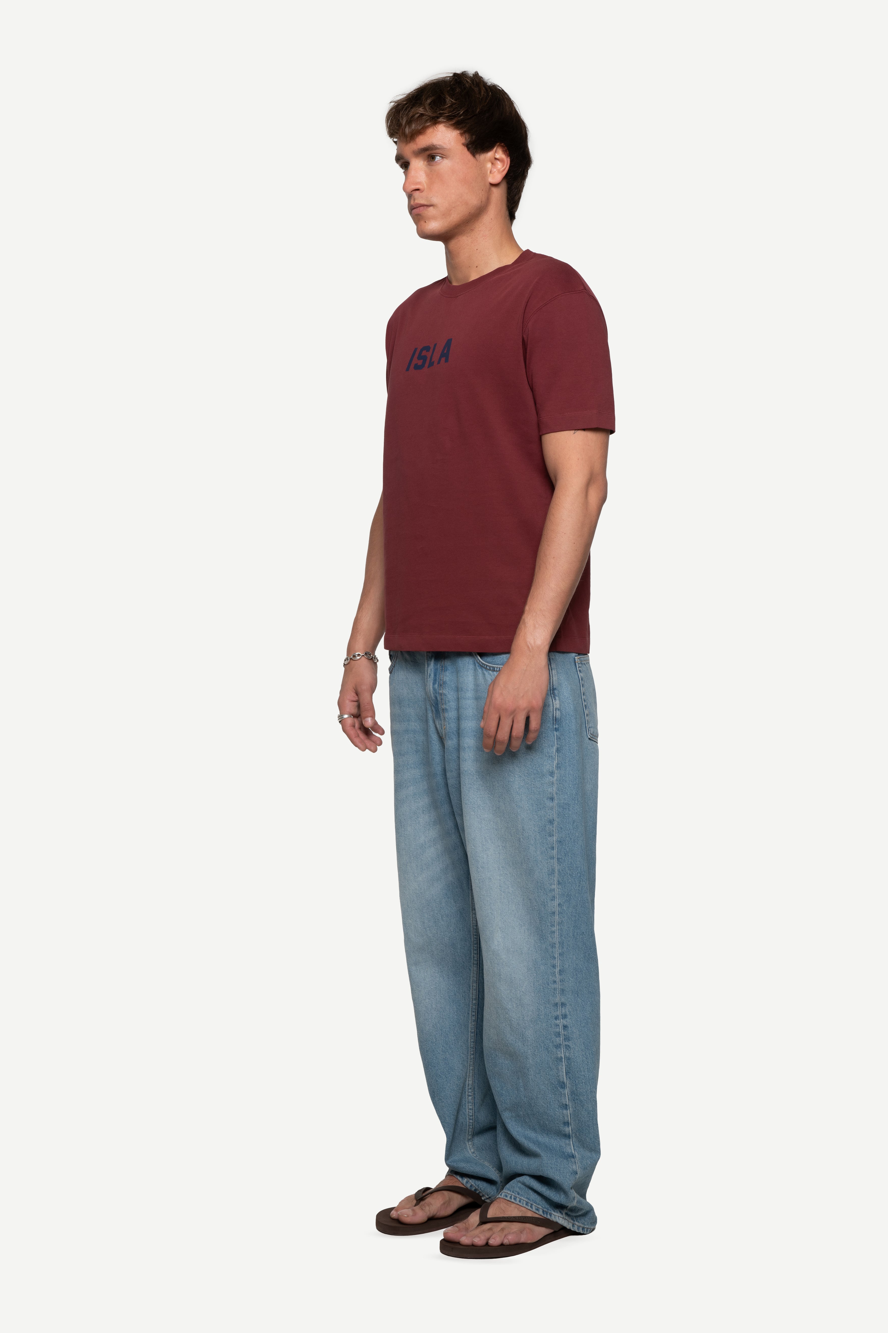 side view of man wearing a regular fit short-sleeved burgundy t-shirt featuring a tonal 'demôssa' logo print on the chest with blue jeans on a white background