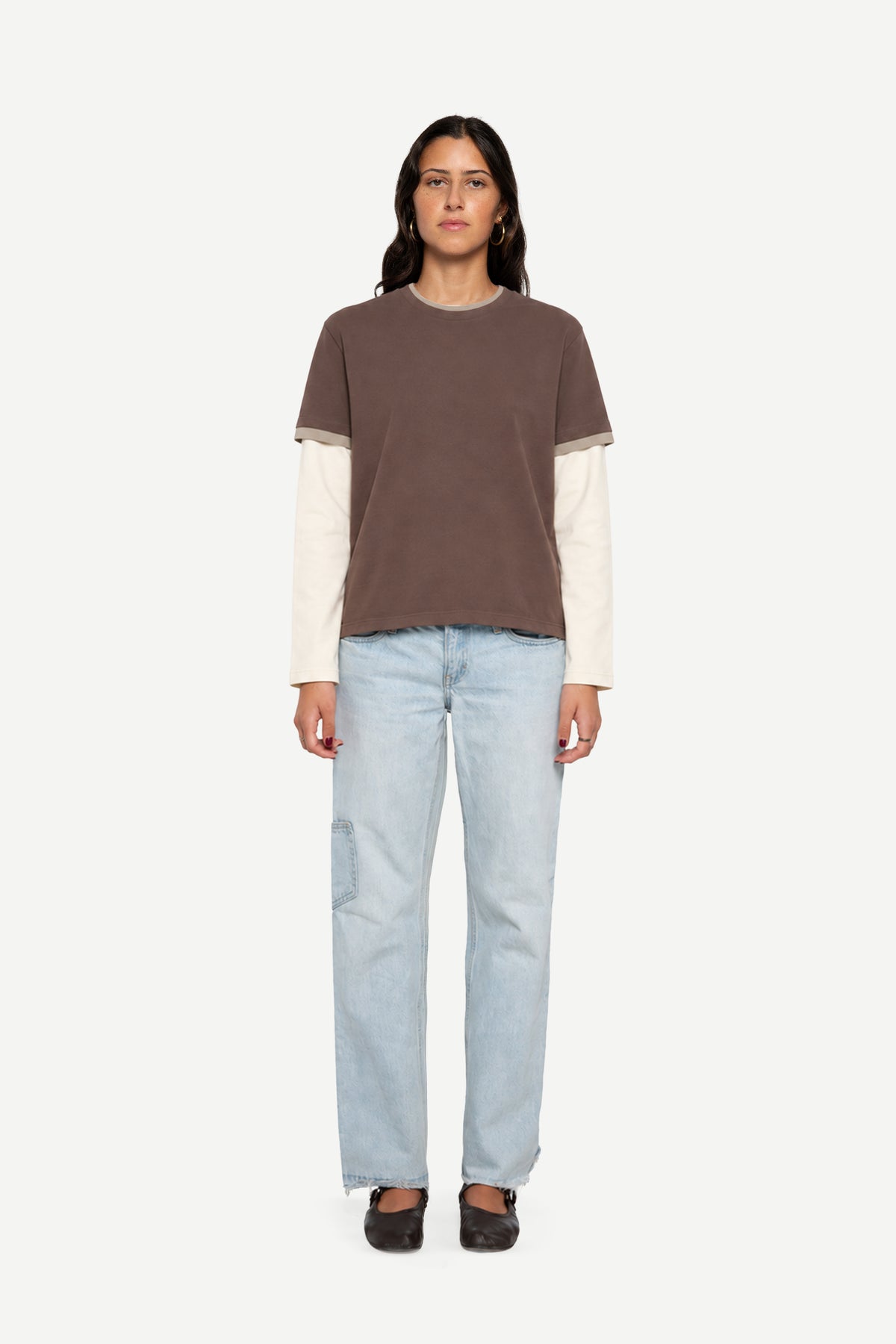 A model stands against a light gray background wearing a color-blocked knit top with a brown upper section and cream sleeves, paired with light-wash blue jeans and dark brown shoes.