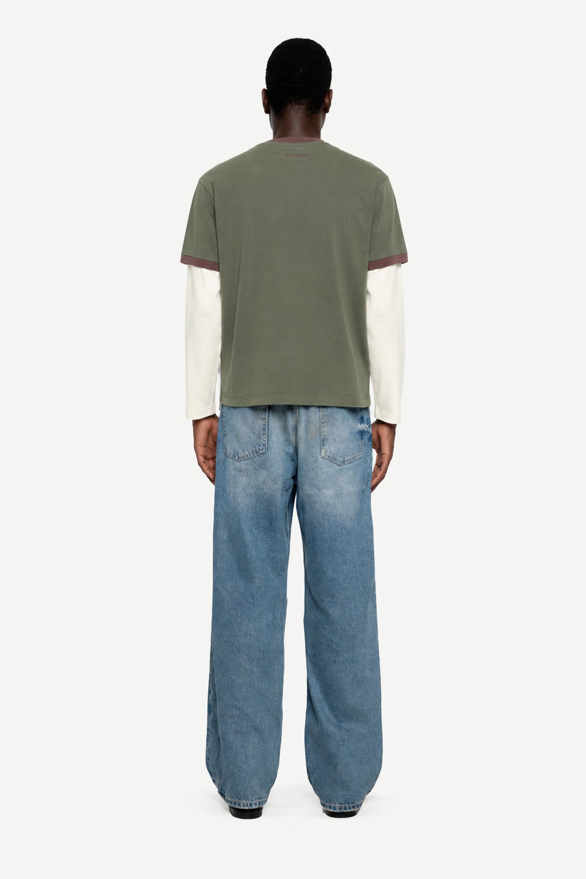 A model viewed from the back wearing a short-sleeve t-shirt layered over a cream long-sleeve shirt, paired with wide-leg blue jeans, against a plain light gray background.