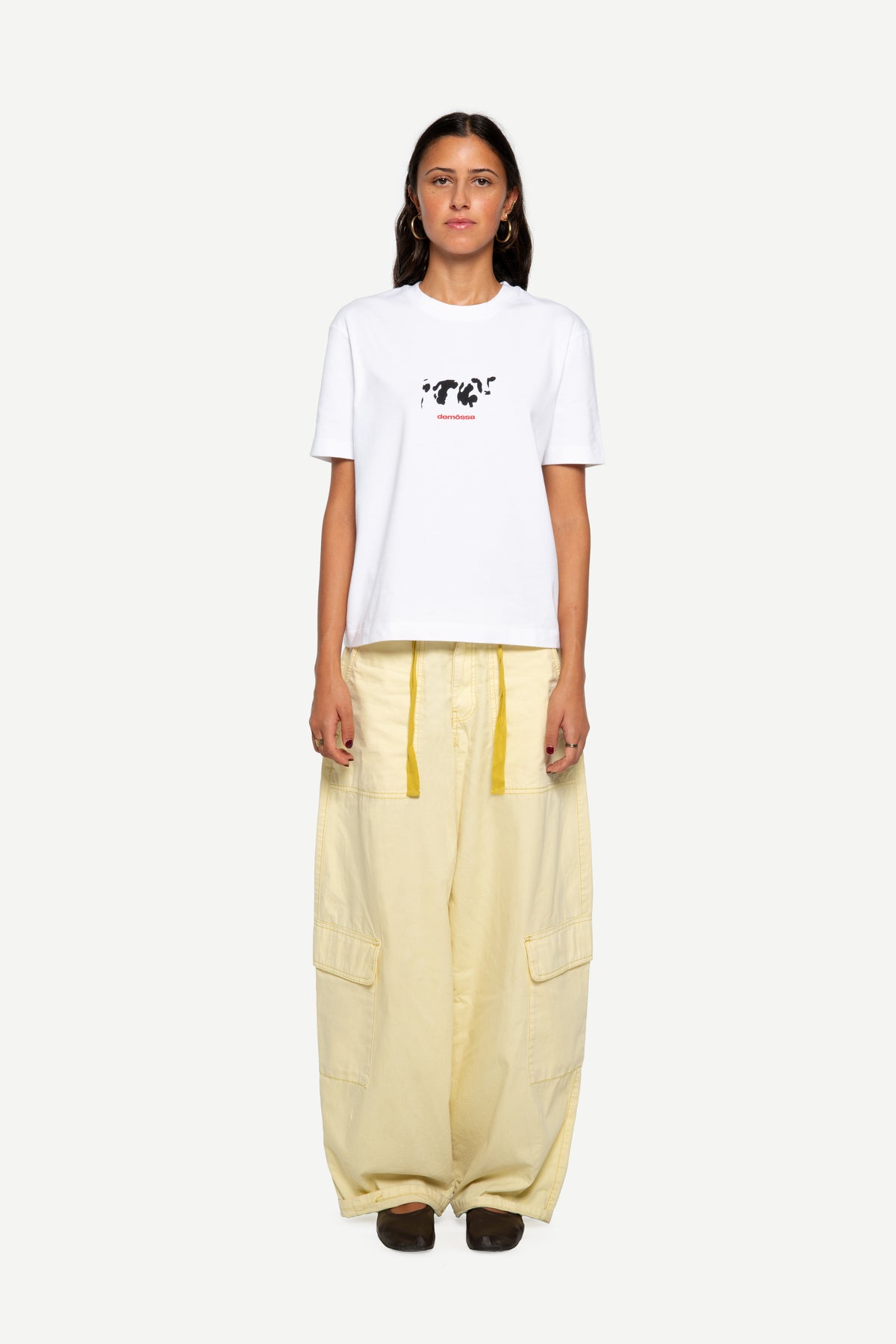 A model stands against a light gray background wearing a white short-sleeve t-shirt with a black graphic and red text on the chest, paired with loose, light yellow cargo pants and dark brown shoes.