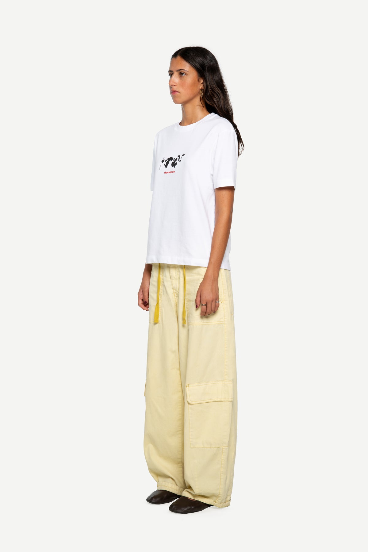 A model is shown from the side wearing a white t-shirt with a small black and red graphic on the chest, paired with wide-leg light yellow cargo pants and dark brown shoes, against a plain light gray background.