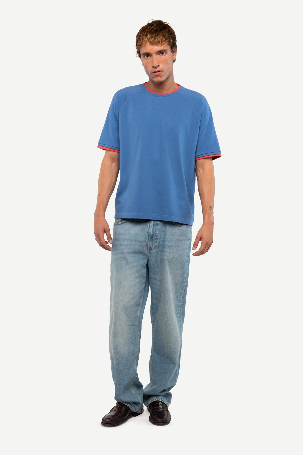 A man modeling a blue short-sleeve t-shirt with red trim on the collar and sleeve cuffs, paired with light blue jeans and dark brown shoes, against a plain light gray background.