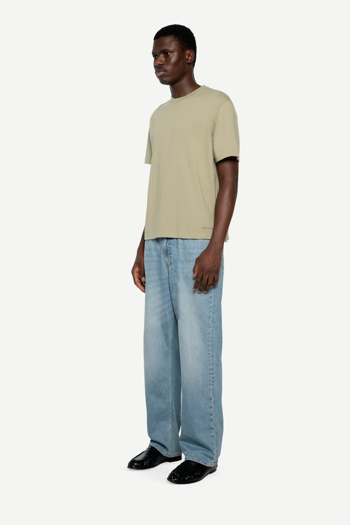 A man wearing a light green short-sleeve t-shirt, wide-leg light blue jeans, and black leather shoes, standing against a plain off-white studio background.