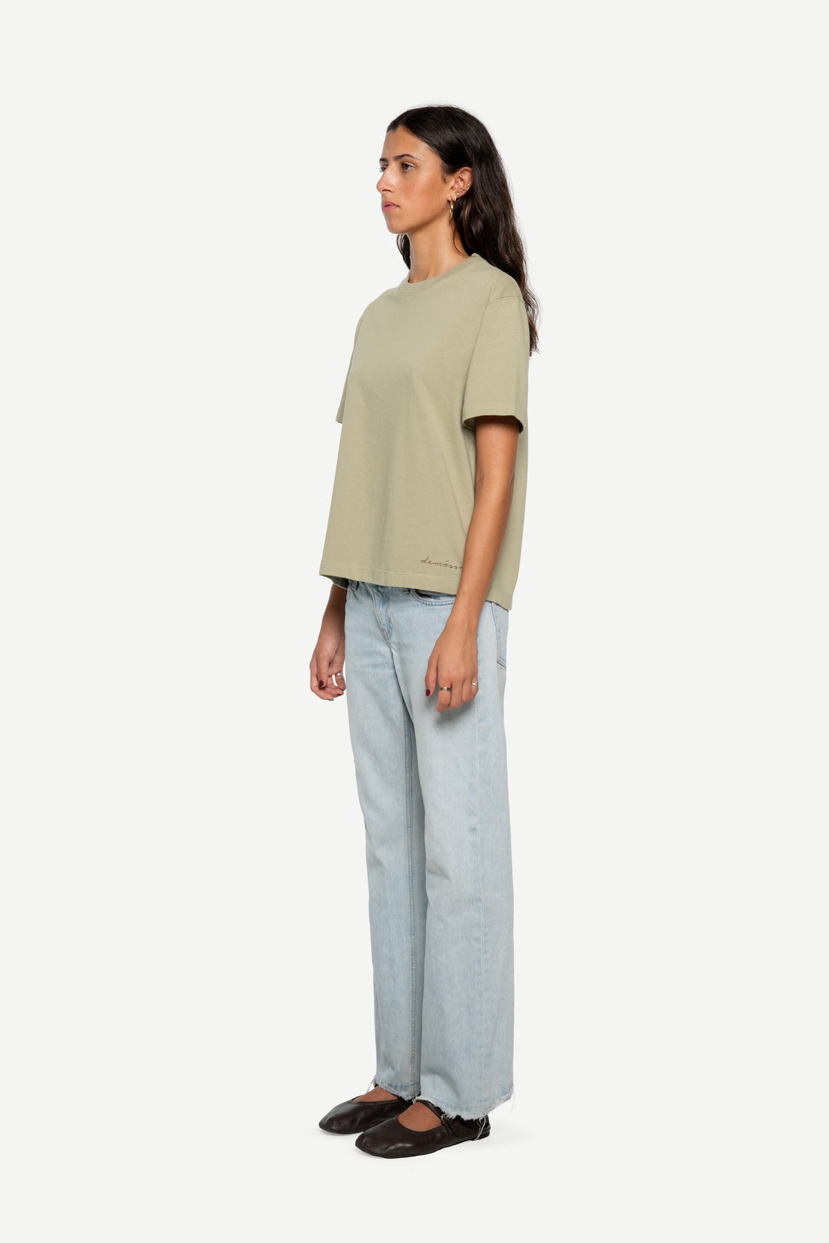 A woman stands in profile wearing a light light green short-sleeve t-shirt, light-wash wide-leg jeans, and dark flat shoes against a plain light gray studio background.