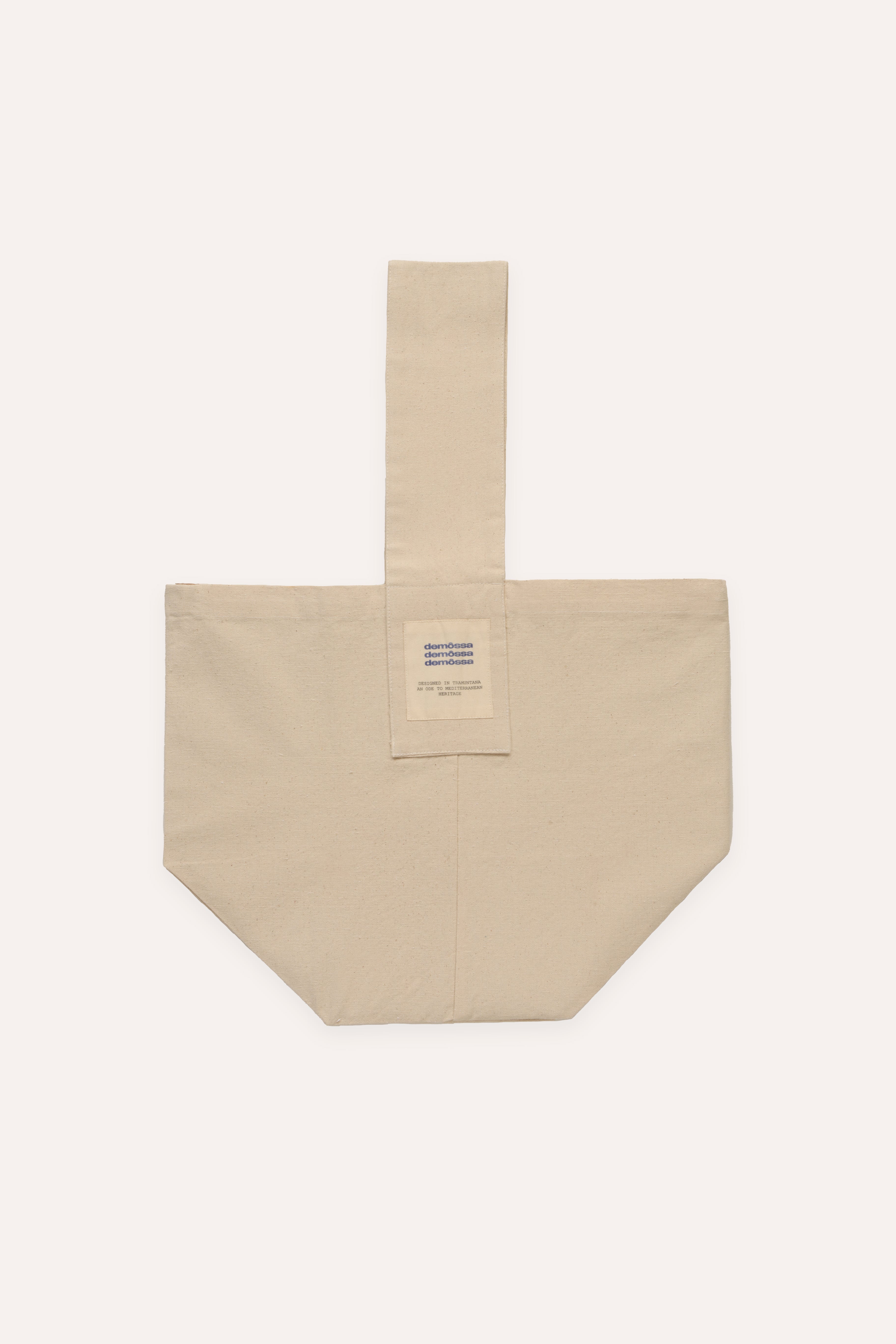 Flat lay of cream canvas tote bag with vertical strap and triple Demossa logo patch on the front