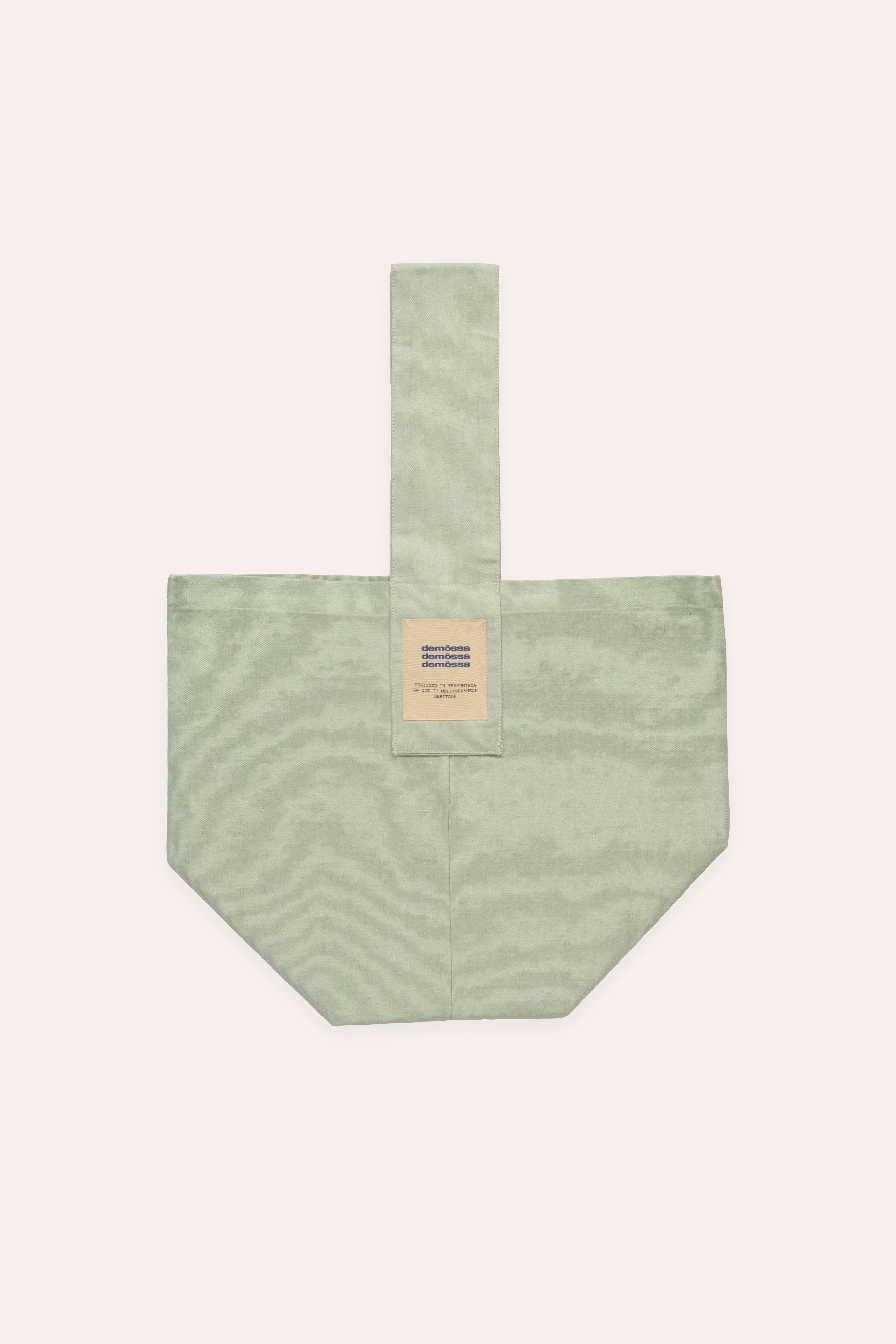 Flat lay of mint green canvas tote bag with central vertical strap and Demossa triple logo patch on front