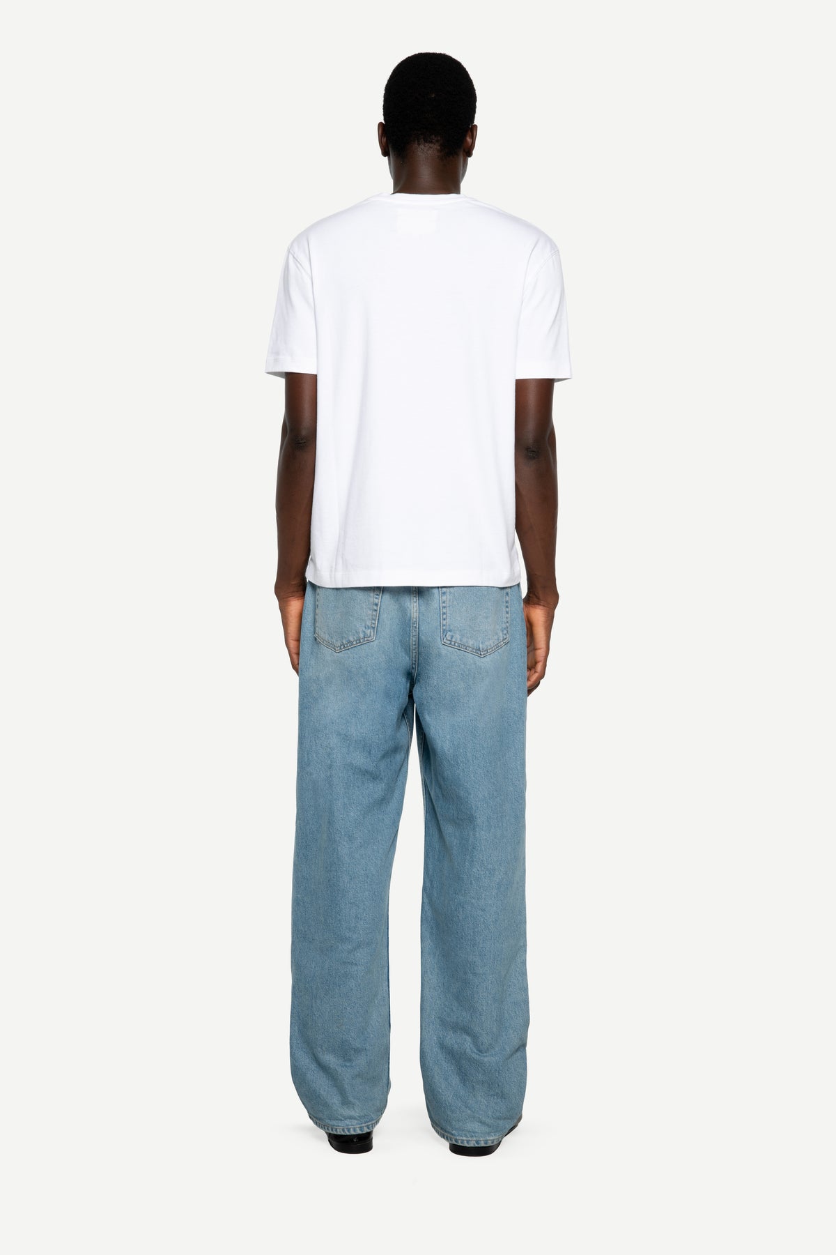 A man viewed from the back wearing a plain white short-sleeve t-shirt and light blue wide-leg jeans, standing against a solid off-white background.