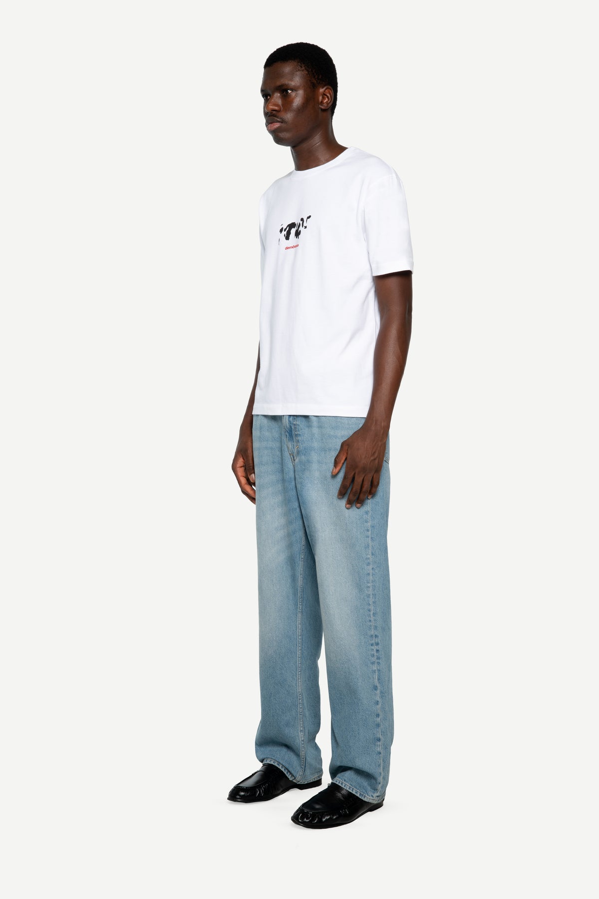 A man modeling a white short-sleeve t-shirt with a small black graphic on the left chest, paired with light-wash wide-leg jeans and black shoes, against a plain light gray background.