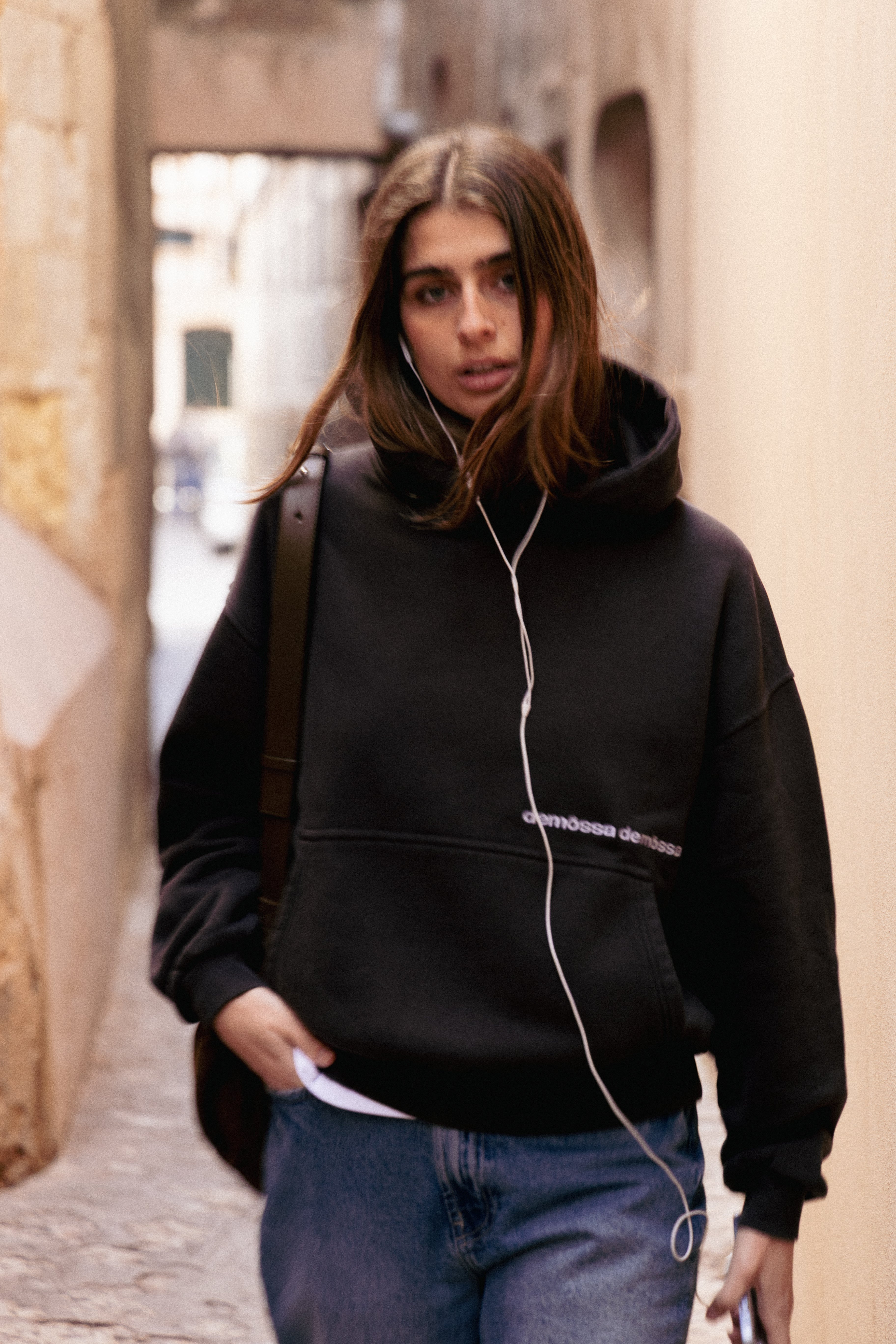 Girl wearing a black hoodie with demôssa logo, cable headphones, walking down a narrow street.