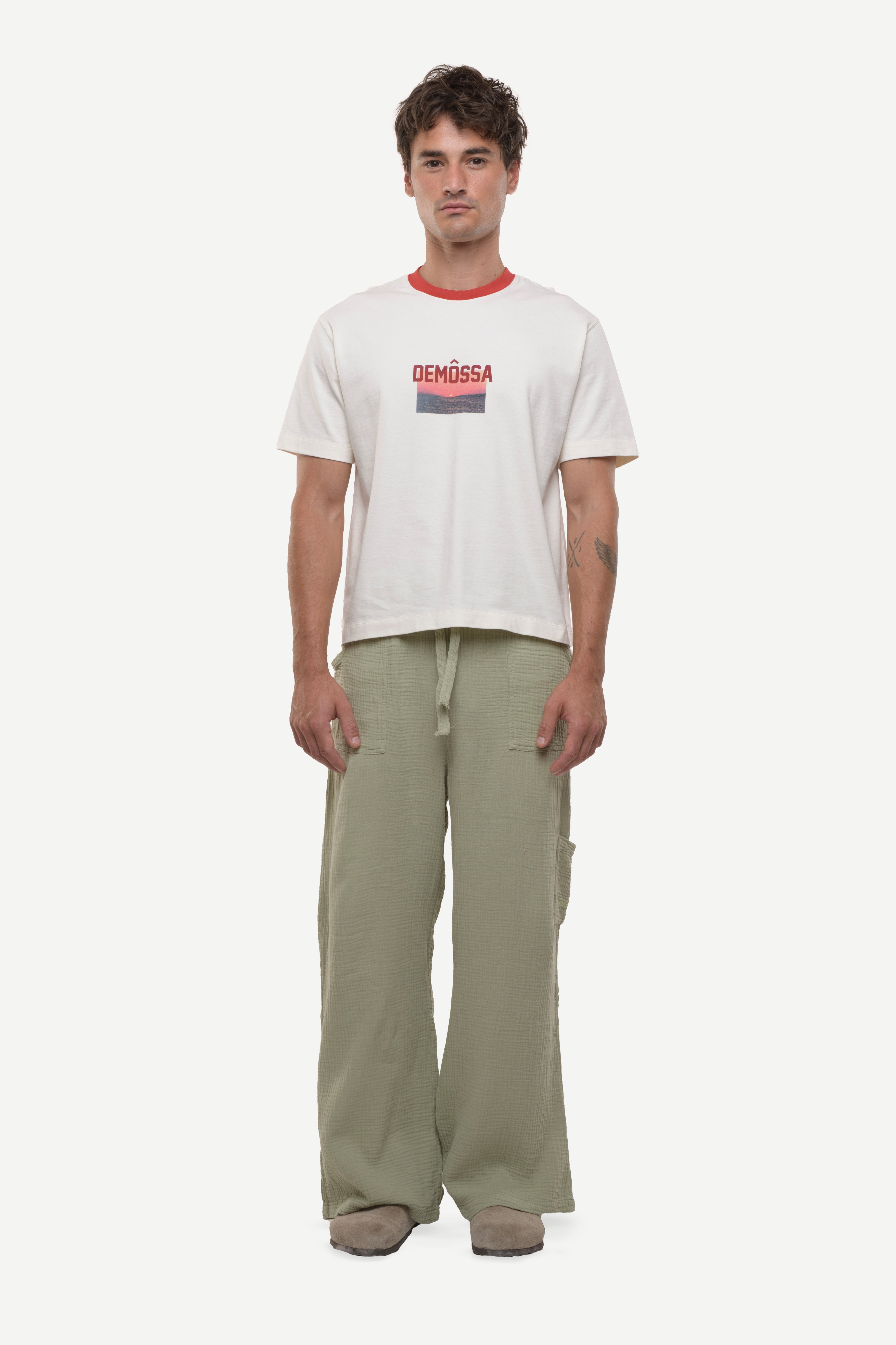 Man wearing a white t-shirt with red demossa logo and sunset picture with green cotton pants white background