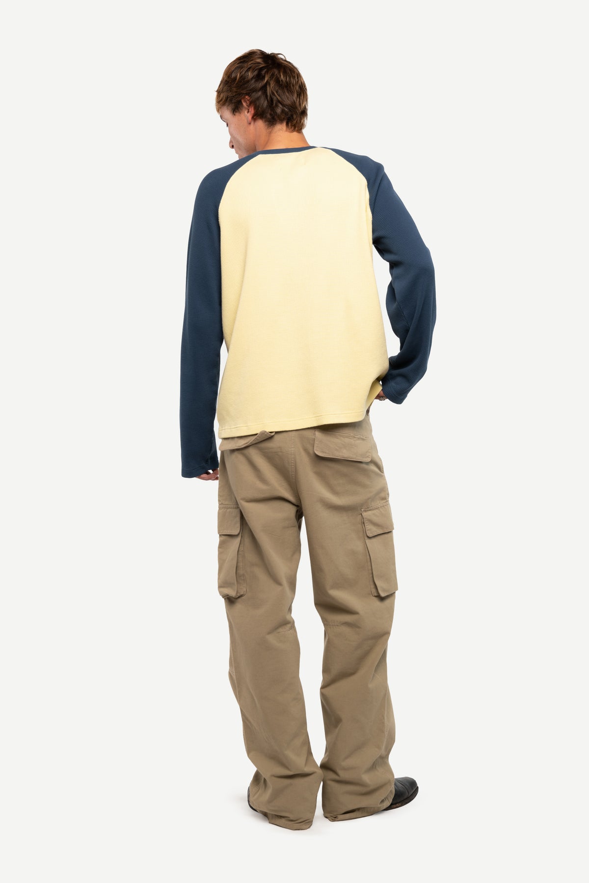 A man shown from the back wearing a raglan-style long-sleeve shirt with a cream body and navy sleeves, paired with tan cargo pants, against a plain light gray studio background.