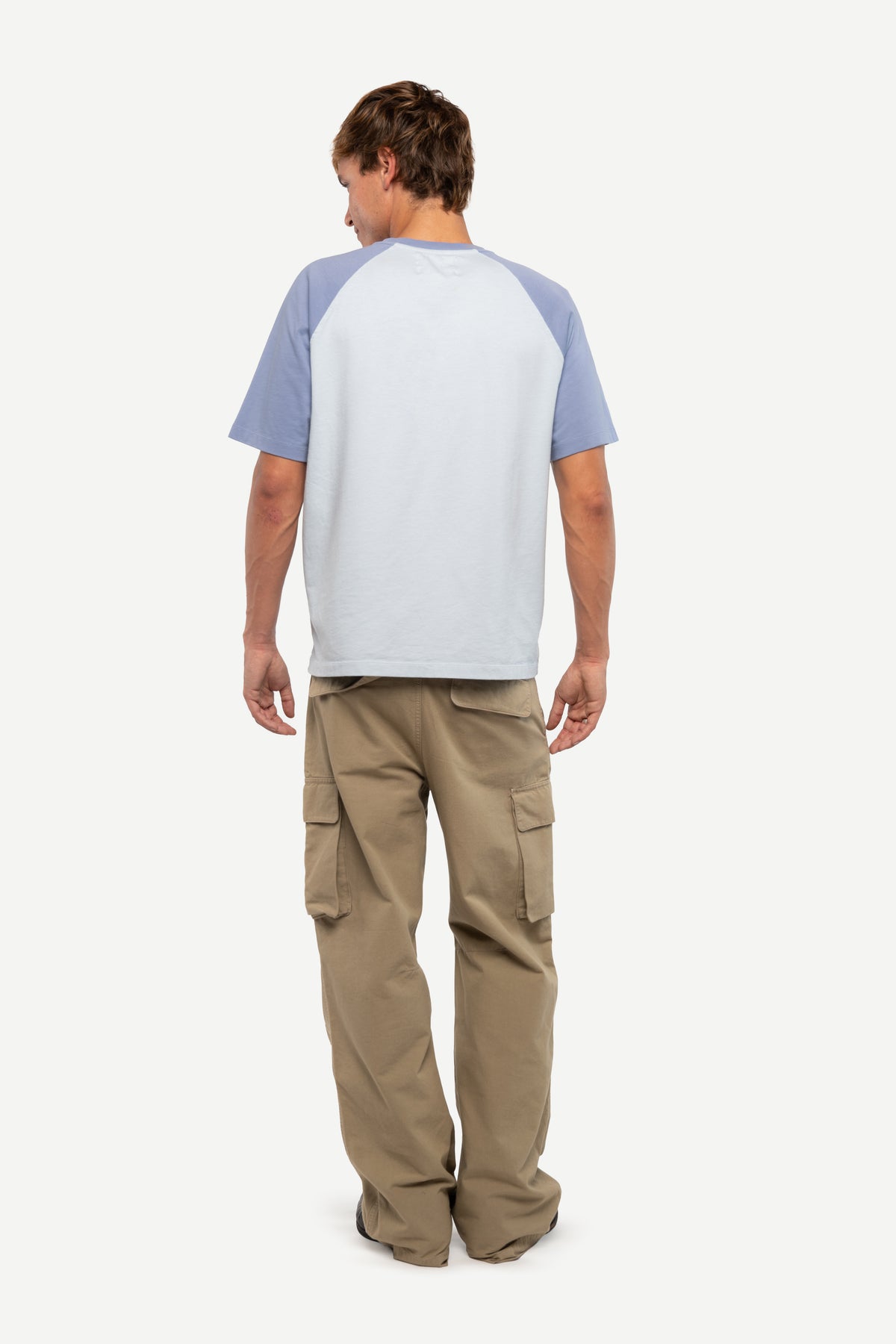 A man viewed from behind wearing a light blue and white raglan t-shirt and tan cargo pants, standing against a solid off-white background.