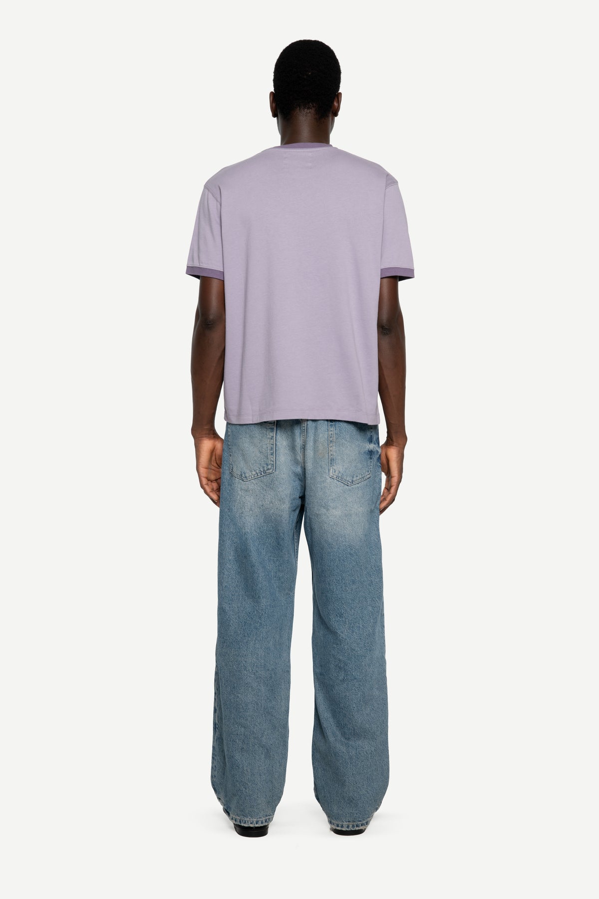 A man viewed from behind wearing a light purple short-sleeve t-shirt and wide-leg blue jeans, standing against a plain light gray background.