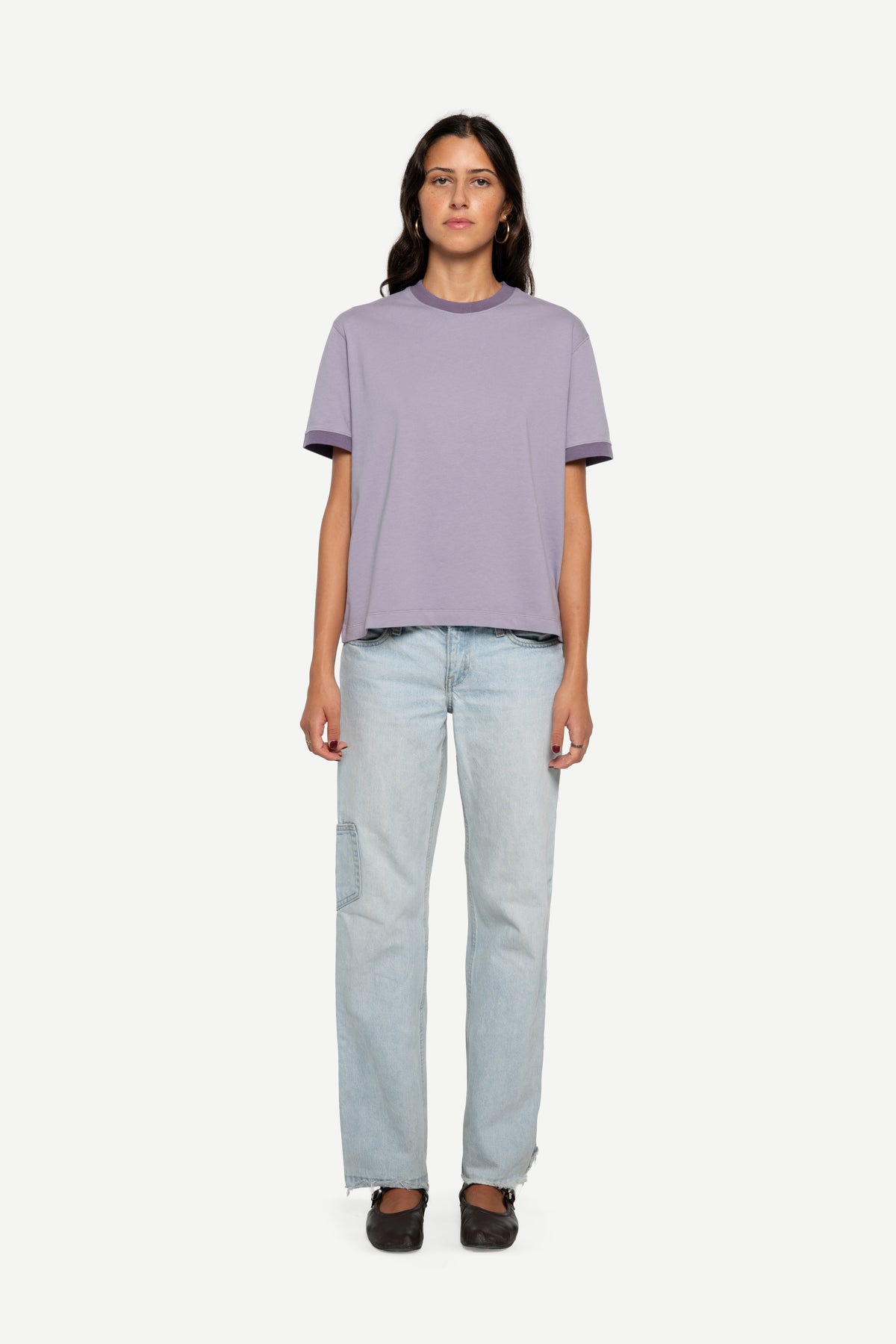 A woman stands against a plain light background wearing a short-sleeve ringer t-shirt in light lavender-purple and light-wash blue jeans.