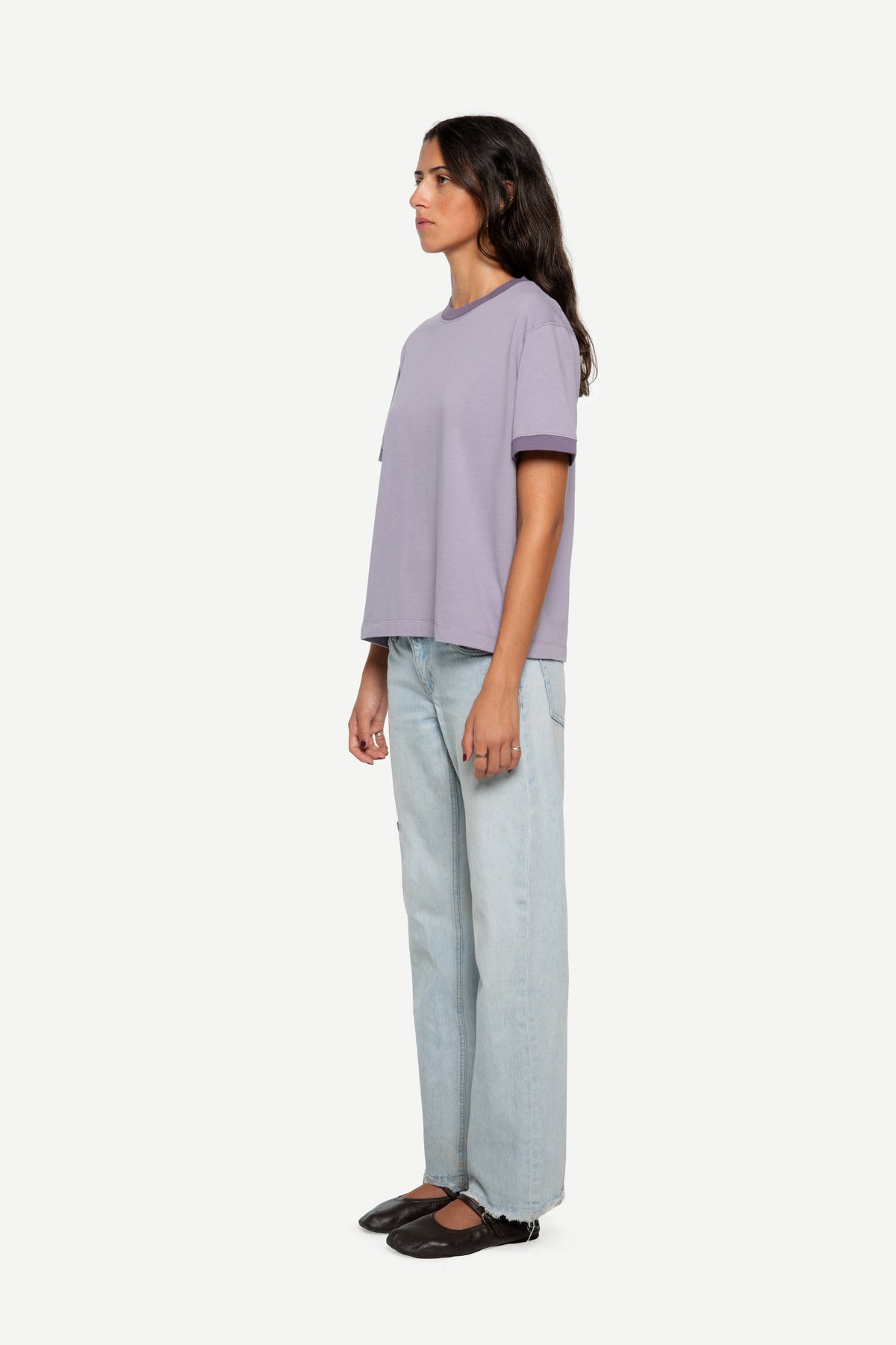 A woman stands in profile wearing a light lavender short-sleeve t-shirt with dark trim, light-wash wide-leg jeans, and dark flat shoes against a plain light gray background.