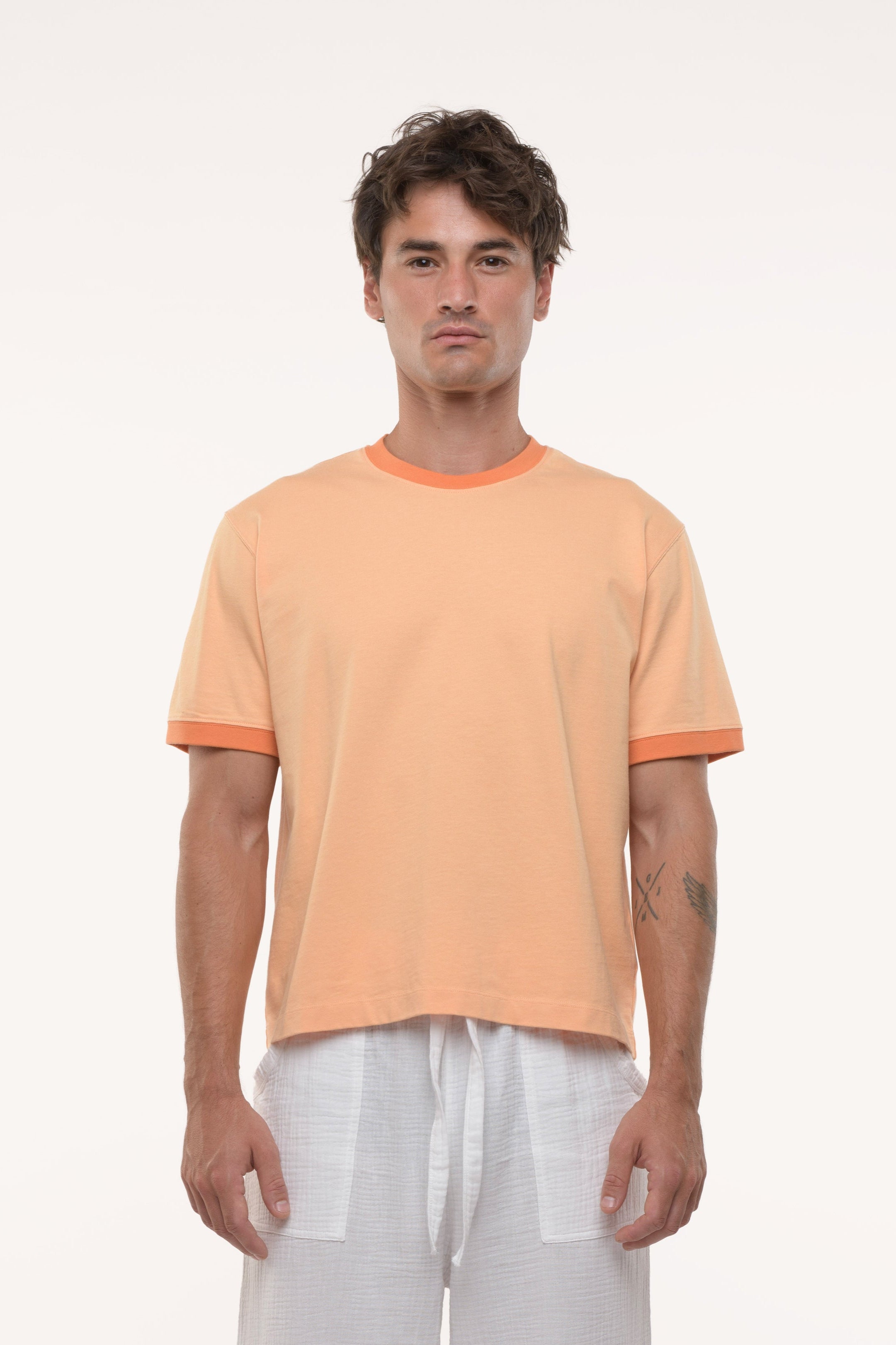 Man wearing an orange t-shirt with contrast collar and sleeve ribs and white pants on a white background