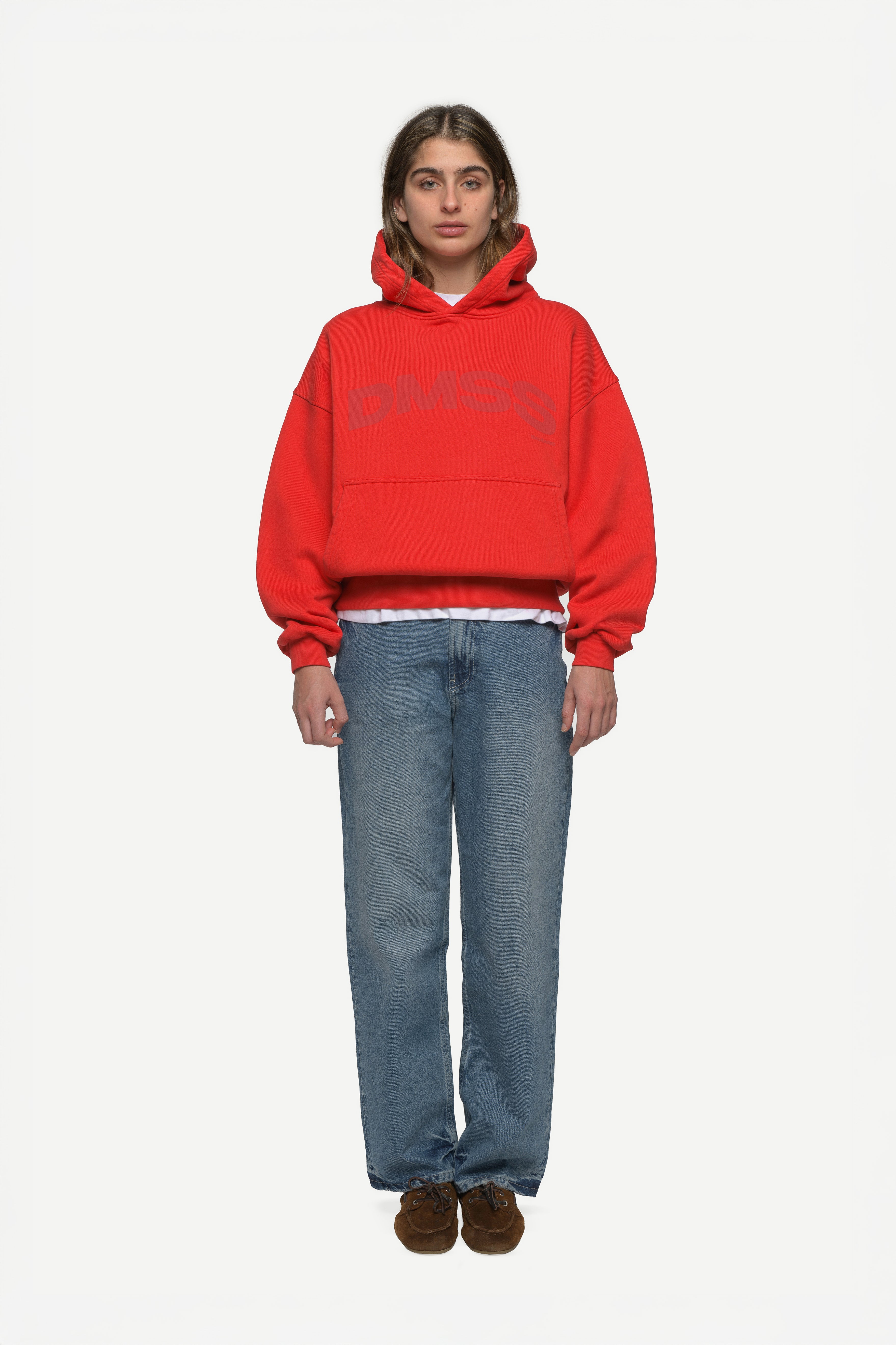 front view of woman wearing a red hoodie with large DMSS logo in a subtle tone and blue jeans on a white background
