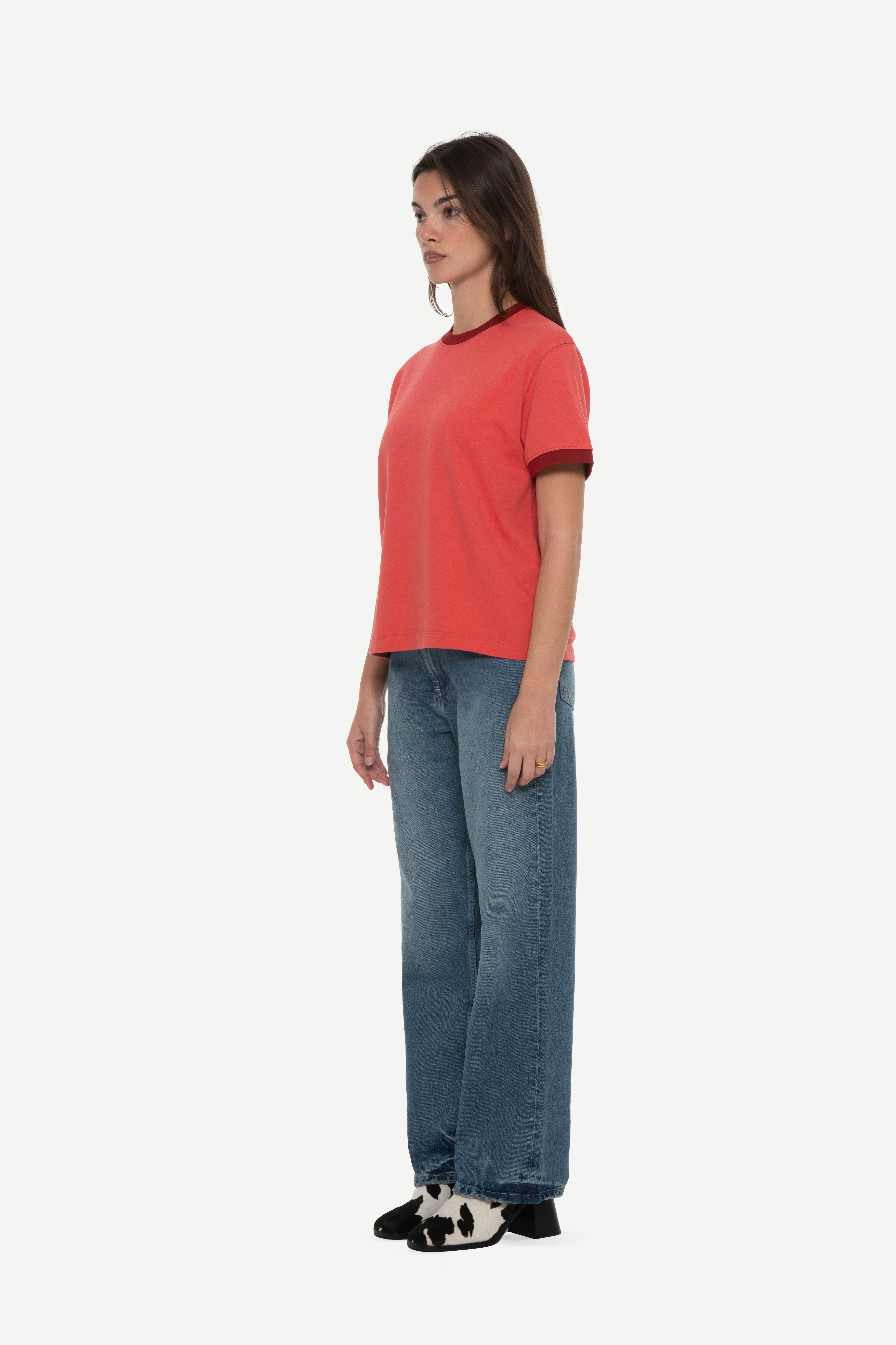 side view of woman wearing a red t-shirt with contrast collar and arm rib and blue jeans on a white background