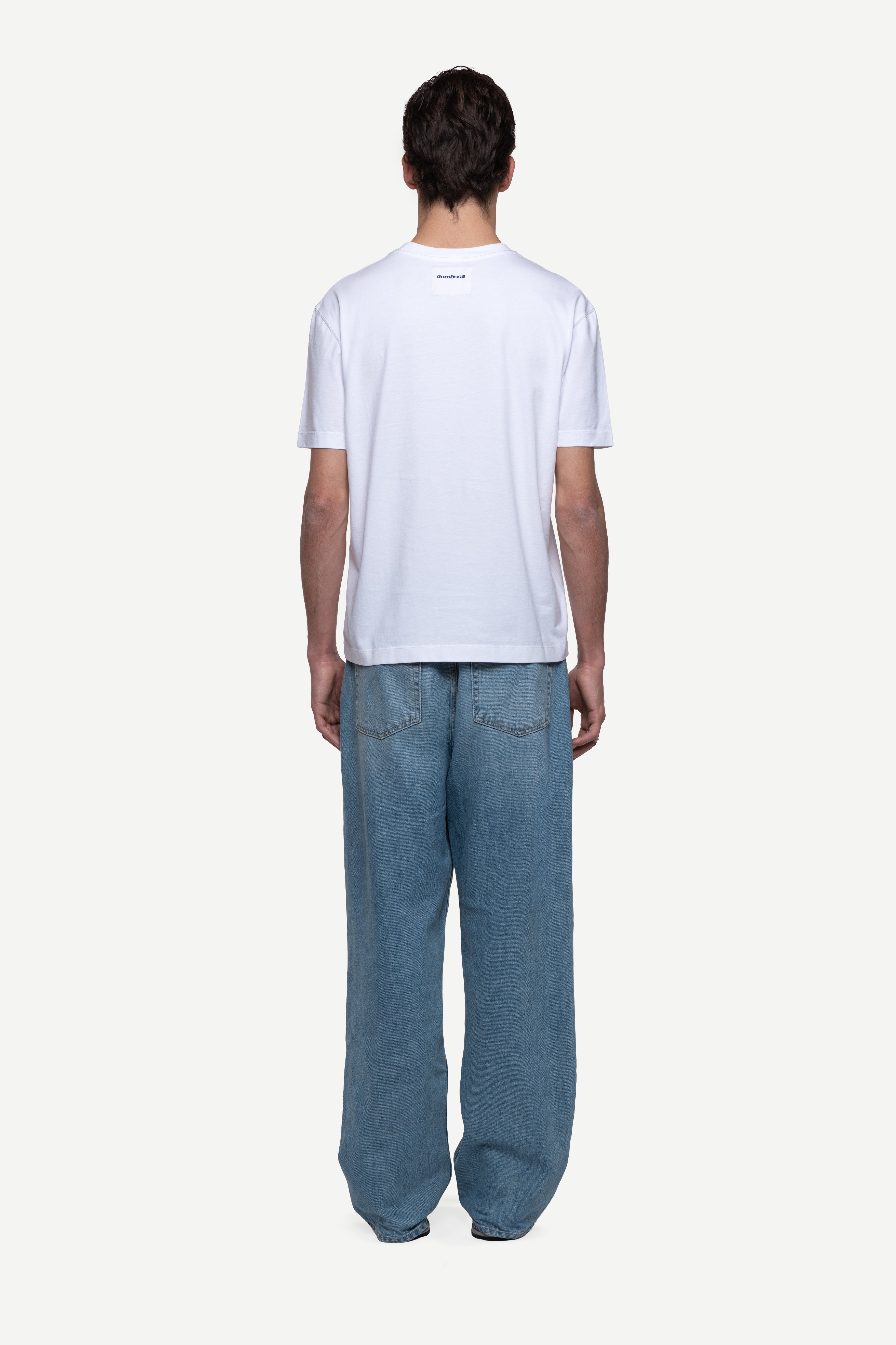 back view of man wearing a regular fit black short-sleeved t-shirt, blue jeans and flip flops on a white background