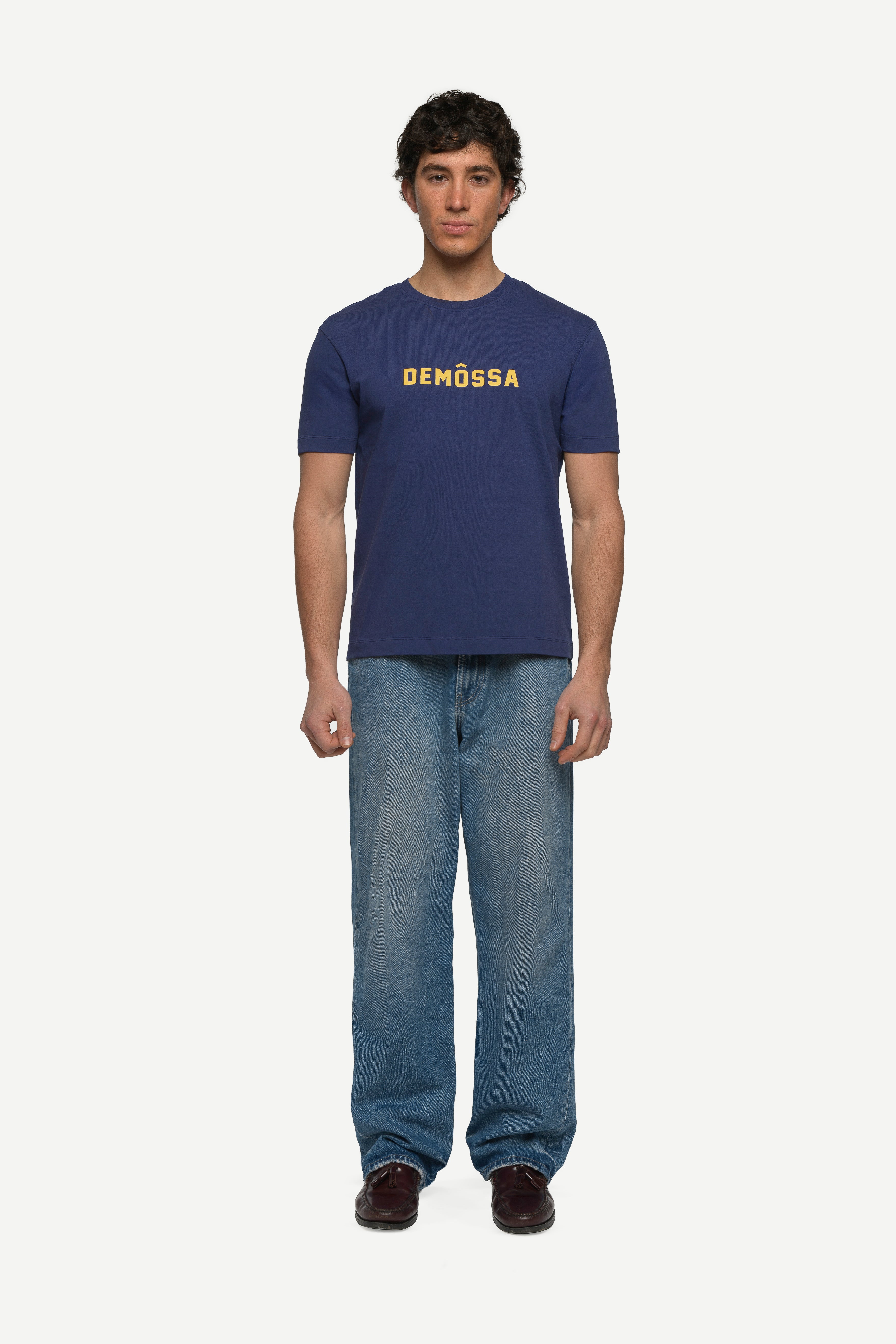 front view of man wearing a navy t-shirt with yellow demossa logo on the chest and blue jeans on a white background