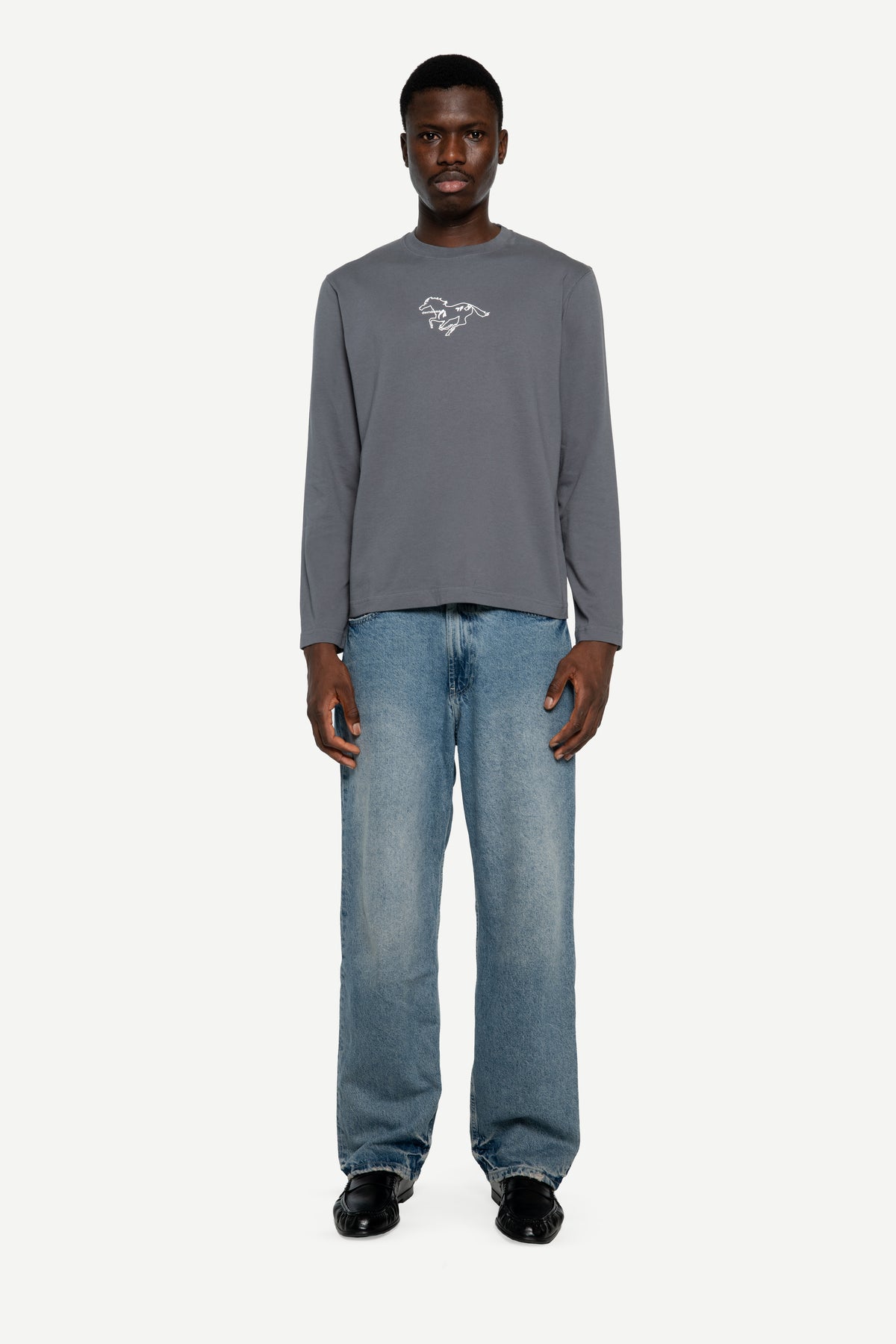 A man modeling a gray long-sleeve t-shirt with a small white graphic on the left chest, paired with light-wash blue jeans and black shoes, against a plain light gray background.
