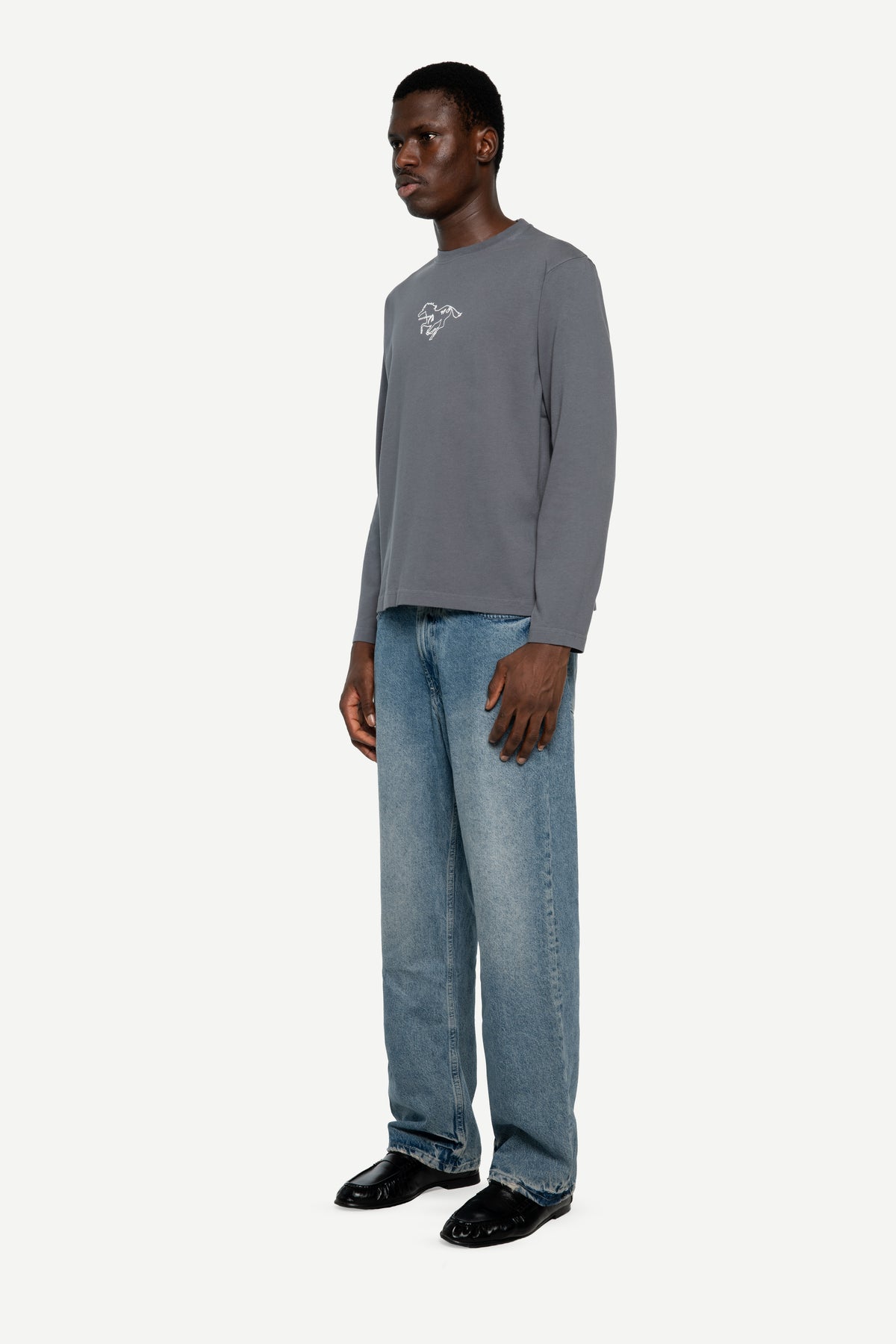 A man wearing a gray long-sleeve crewneck sweatshirt with a small white embroidered logo on the left chest, paired with light blue jeans and black loafers, standing against a light gray studio background.