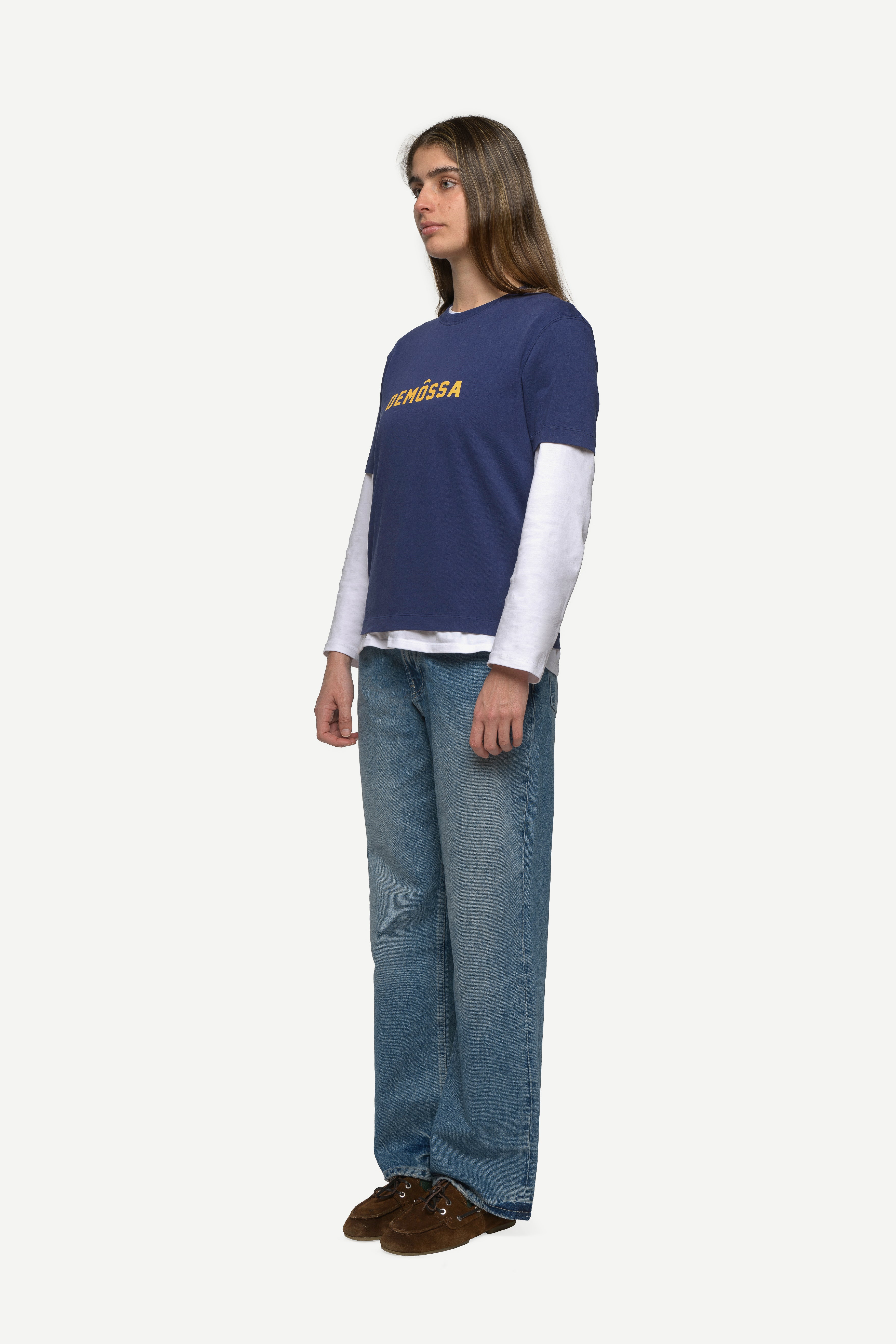 side view of woman wearing a navy t-shirt with yellow demossa logo on the chest and blue jeans on a white background