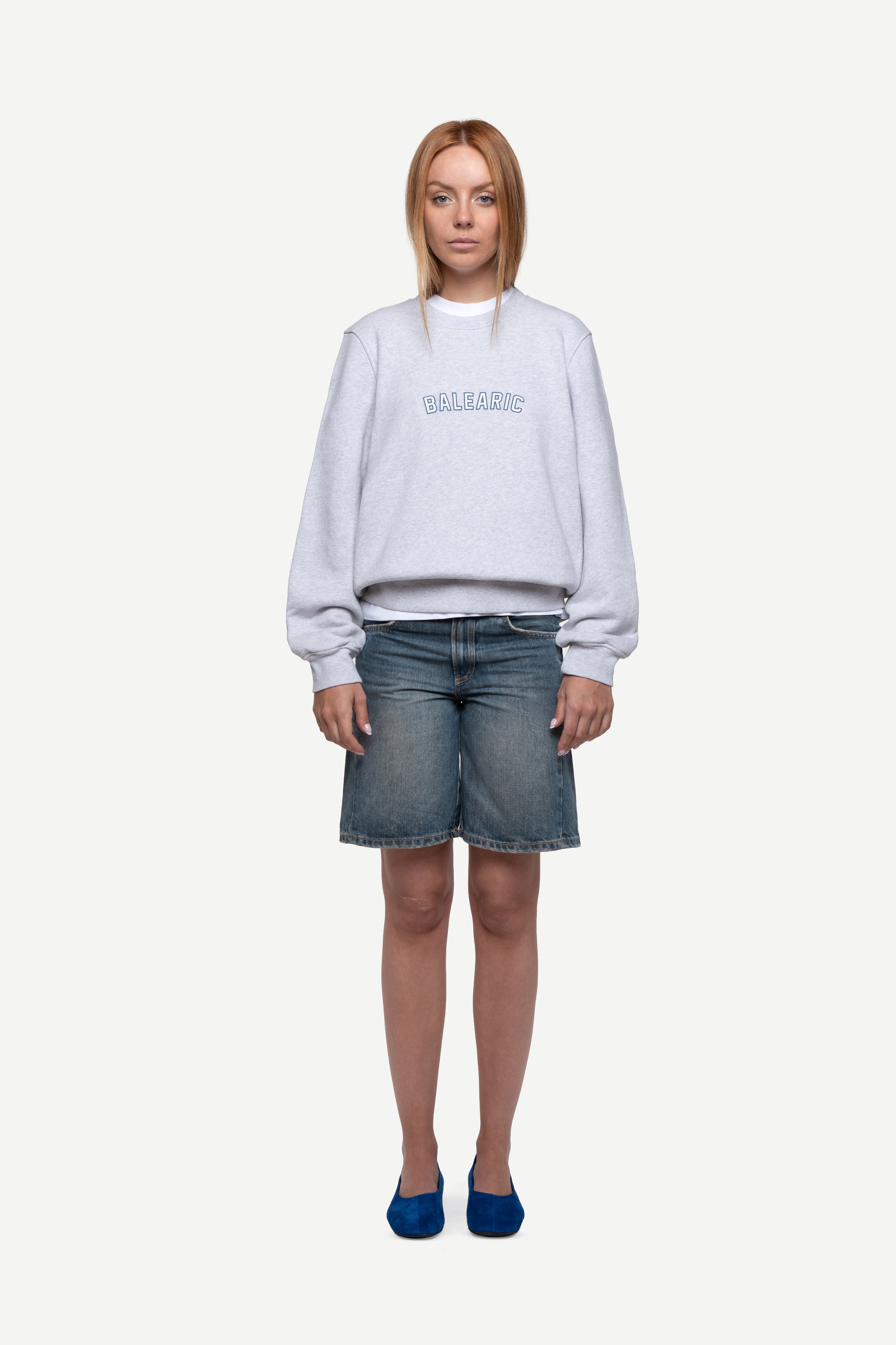 front view of woman wearing a melange grey relaxed-fit sweatshirt with "Balearic" print on the chest, blue short jeans and ballet flats on a white background