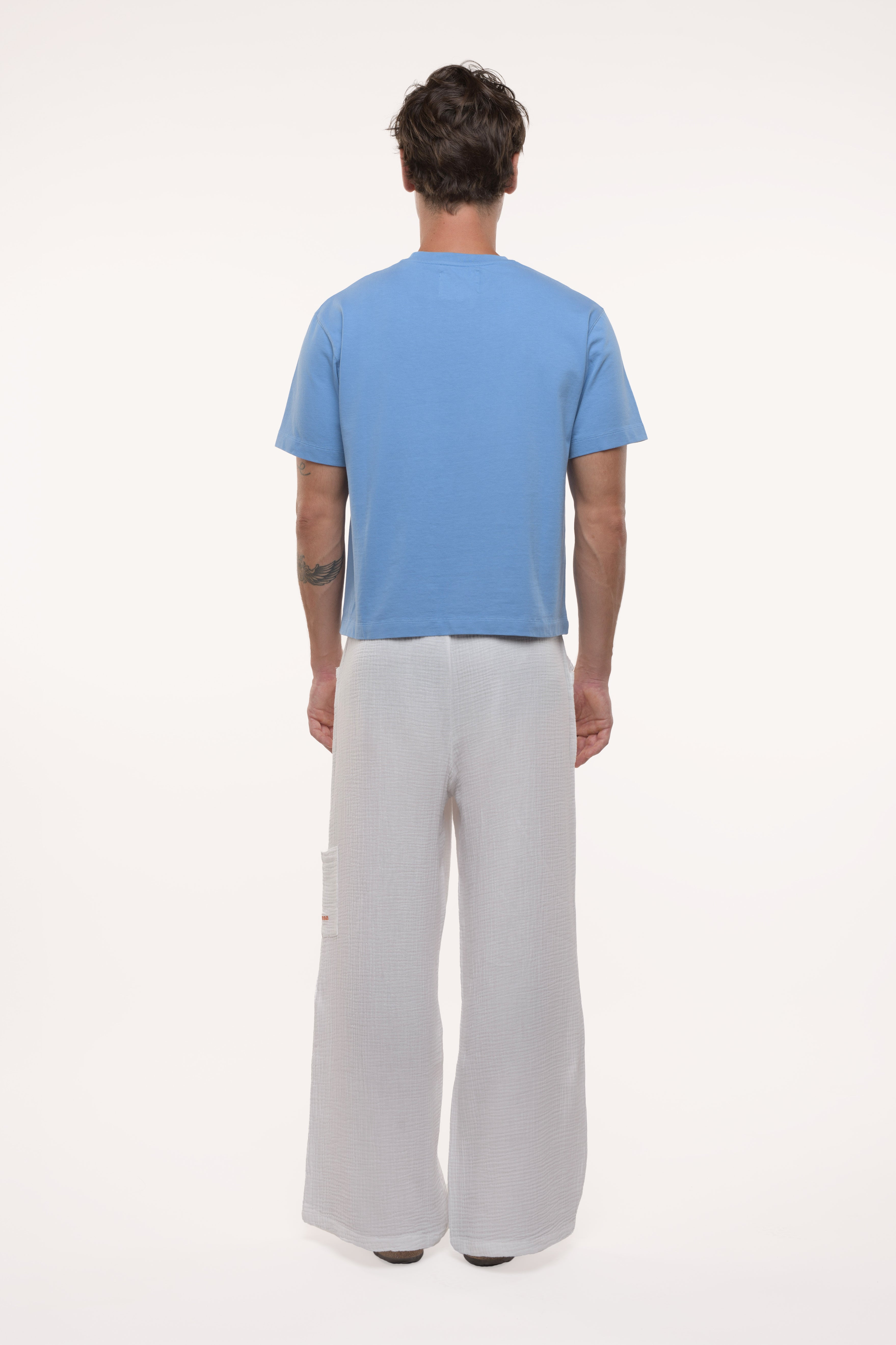 Back view of a man wearing a blue t-shirt and white cotton pants on a white background