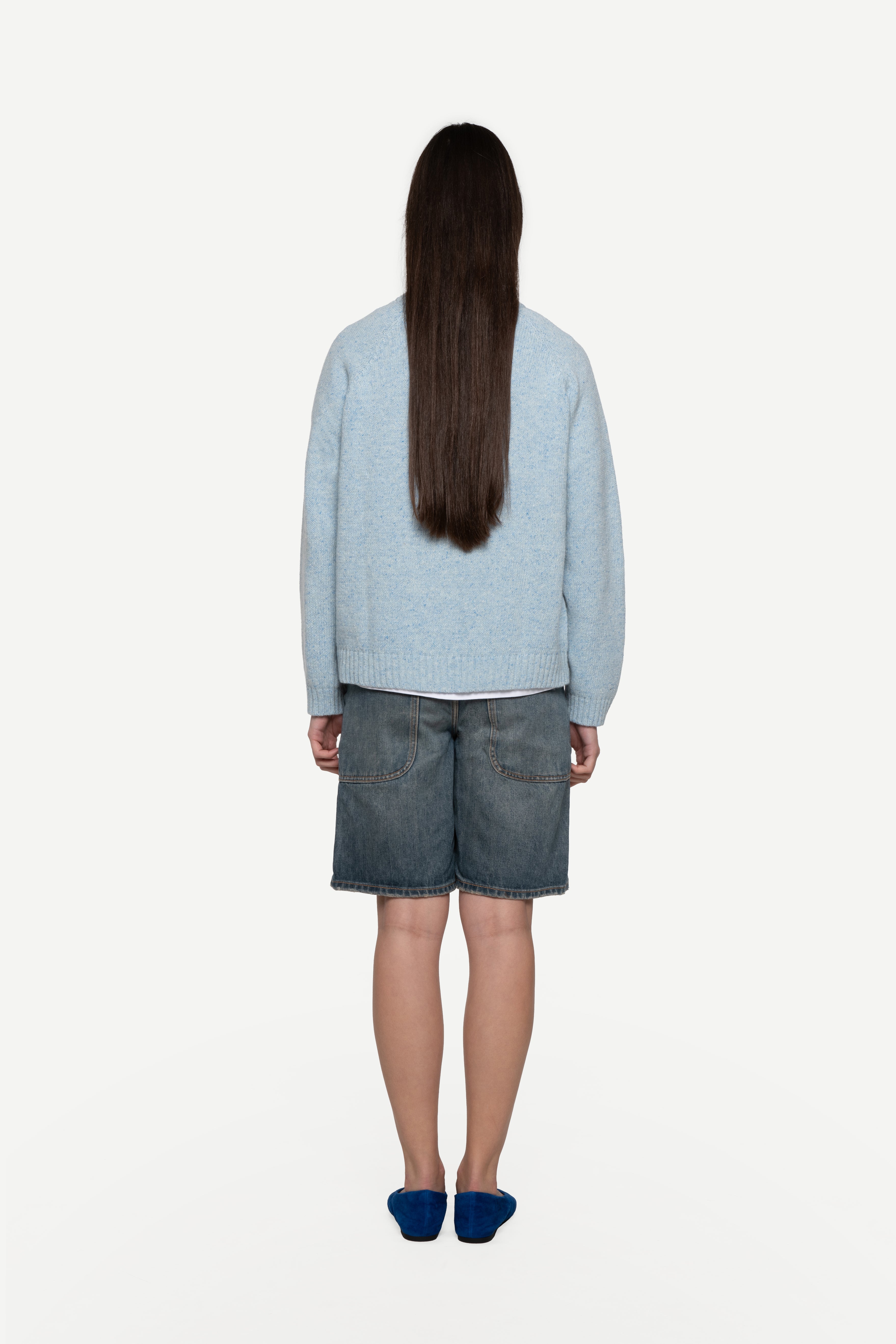 back view of woman wearing a blue sweater made from a soft blend of wool and silk with blue short jeans on a white background