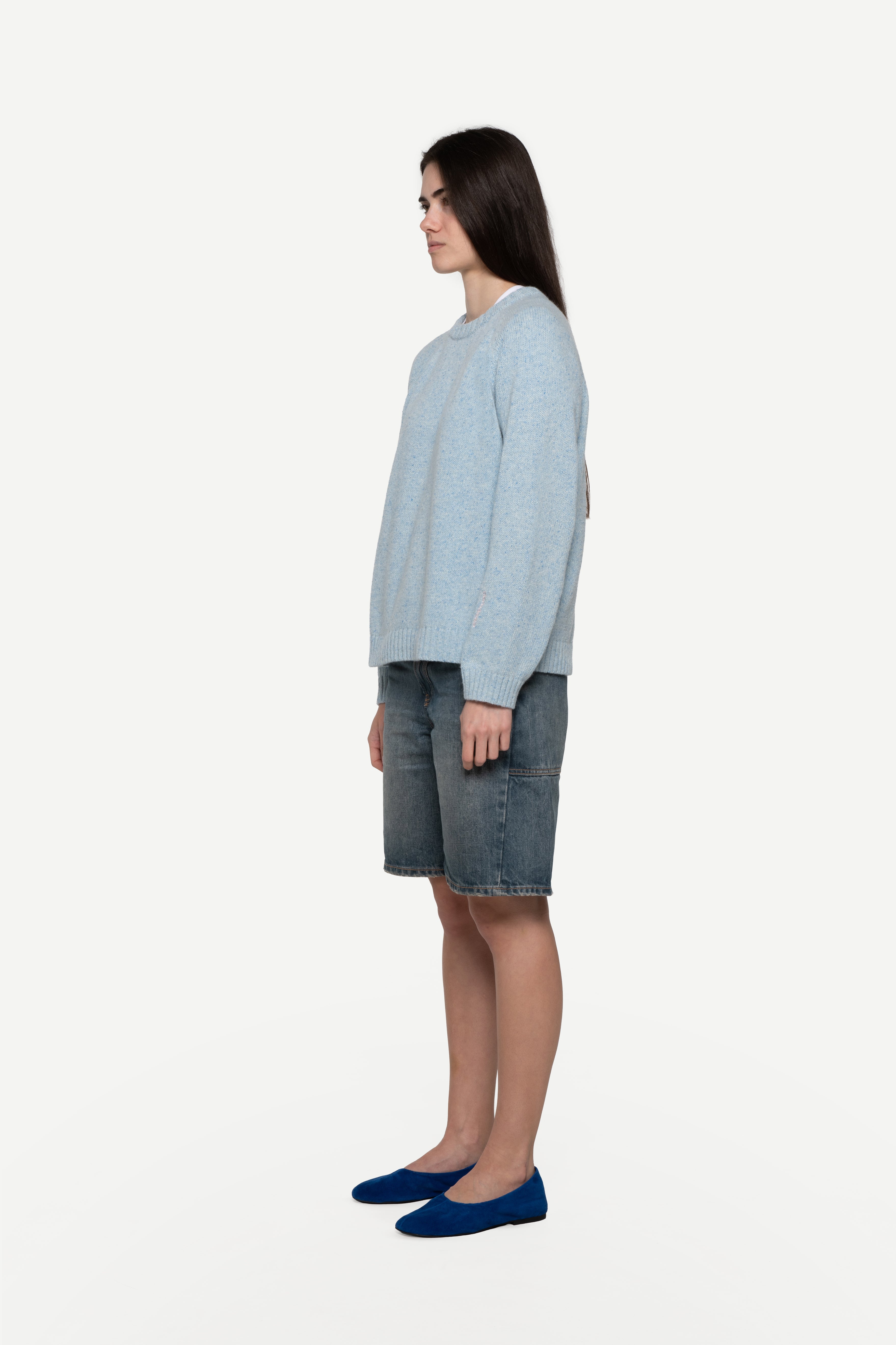 side view of woman wearing a blue sweater made from a soft blend of wool and silk with blue short jeans on a white background