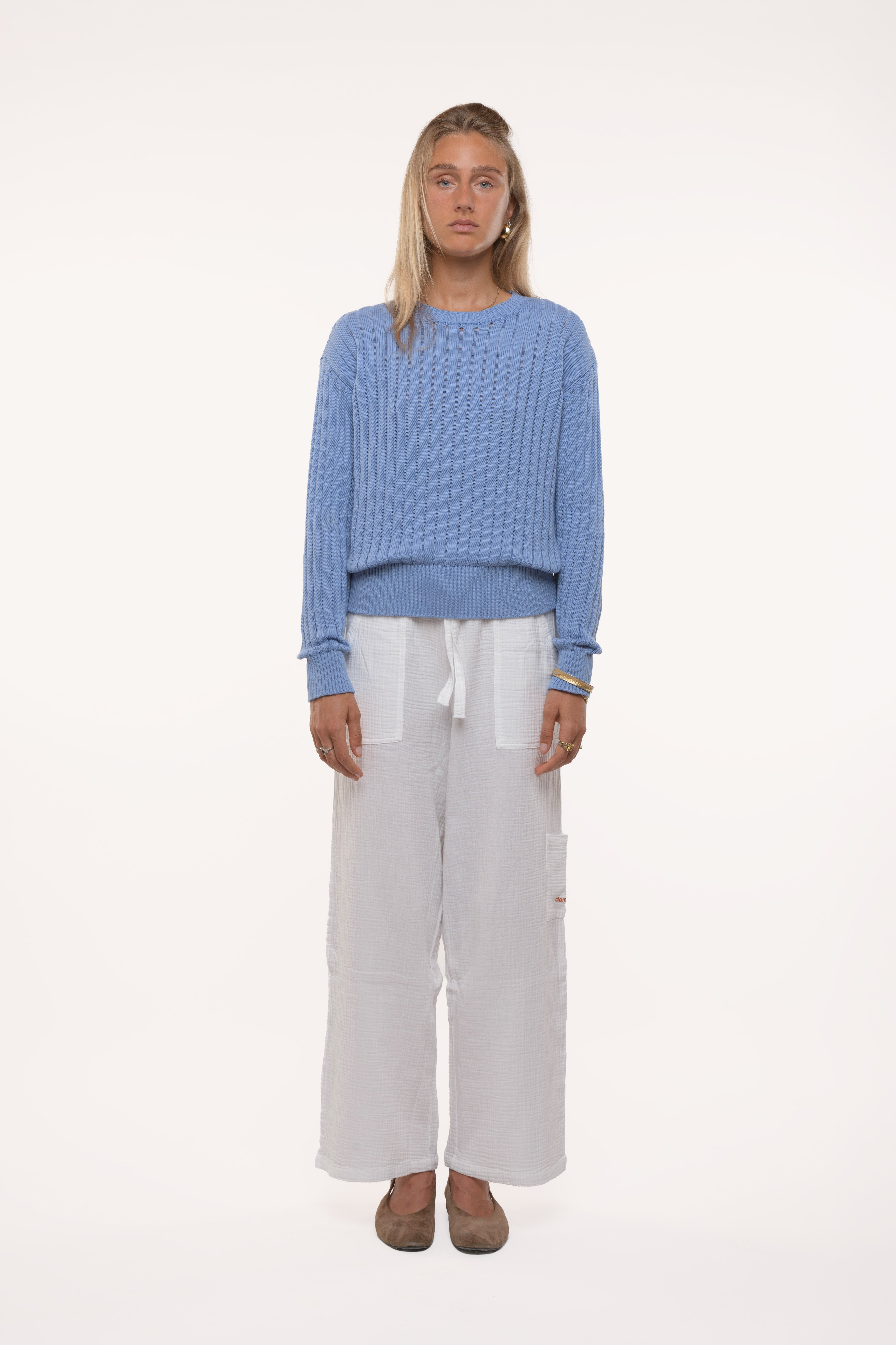 Woman wearing a blue open knit sweater and white pants on a white background